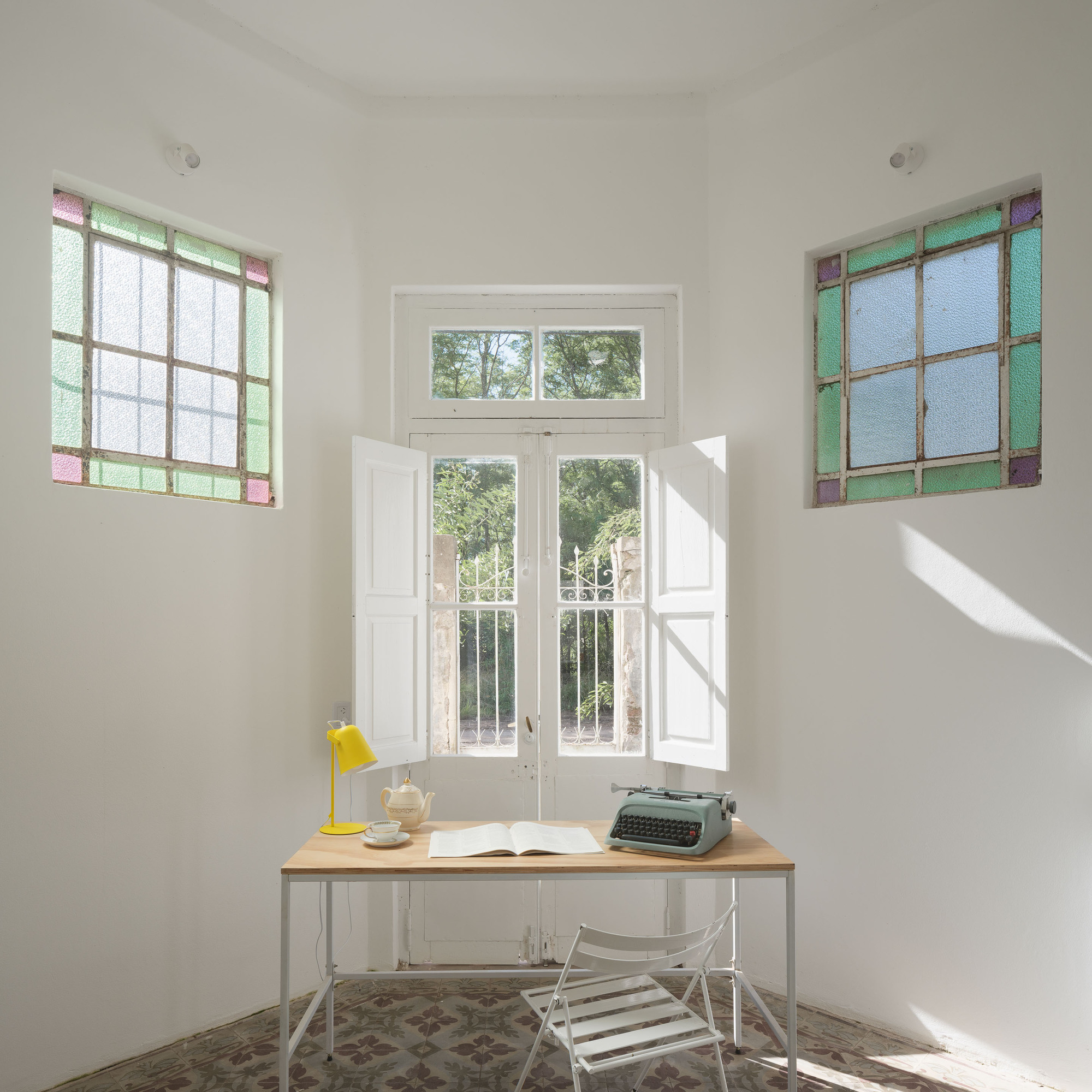 Writing desk beneath French doors flanked by colored glass windows with sunlight streaming across patterned tile floor