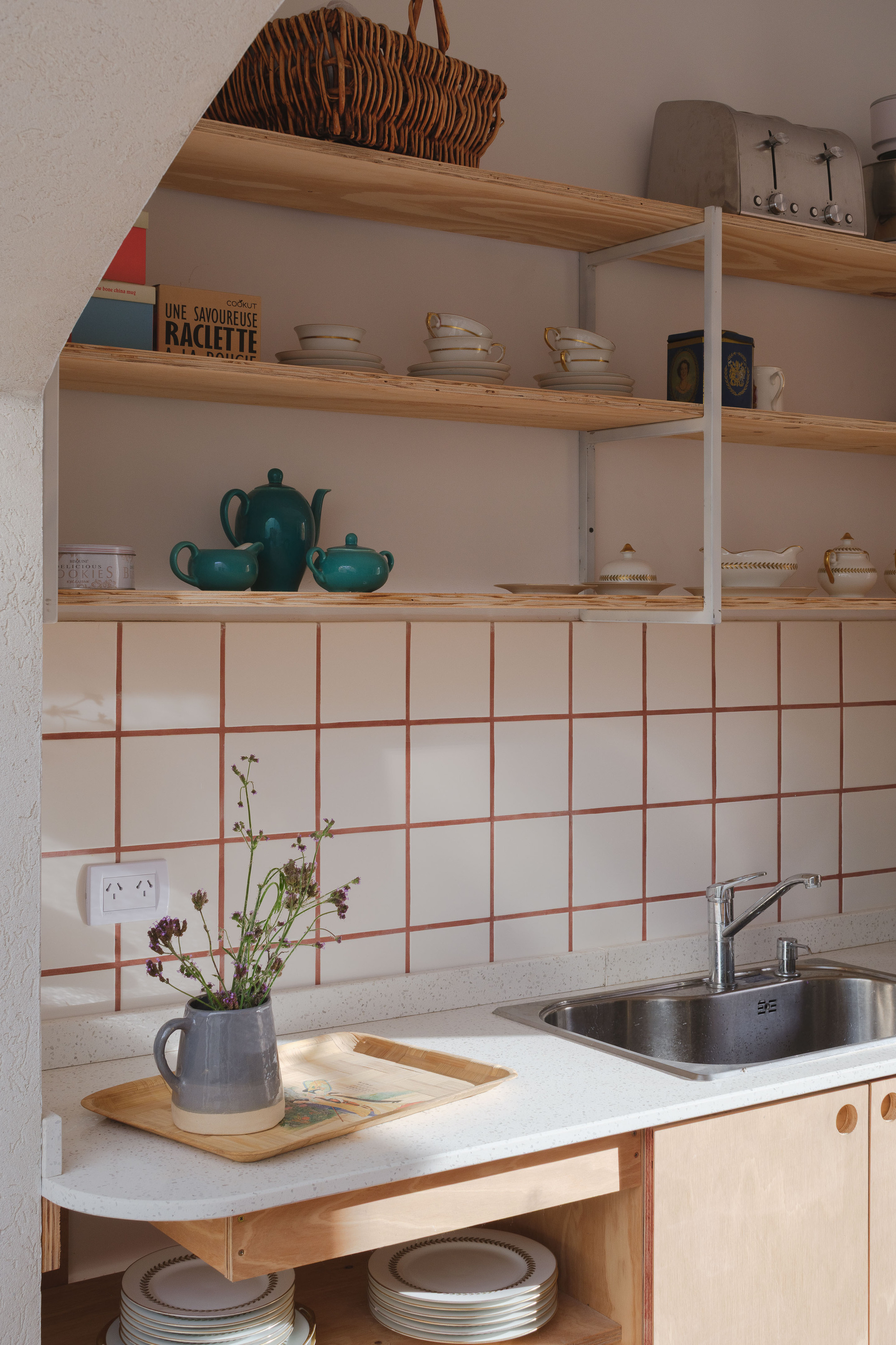 Kitchen corner with white square tile backsplash, timber shelving and a jug of dried wildflowers
