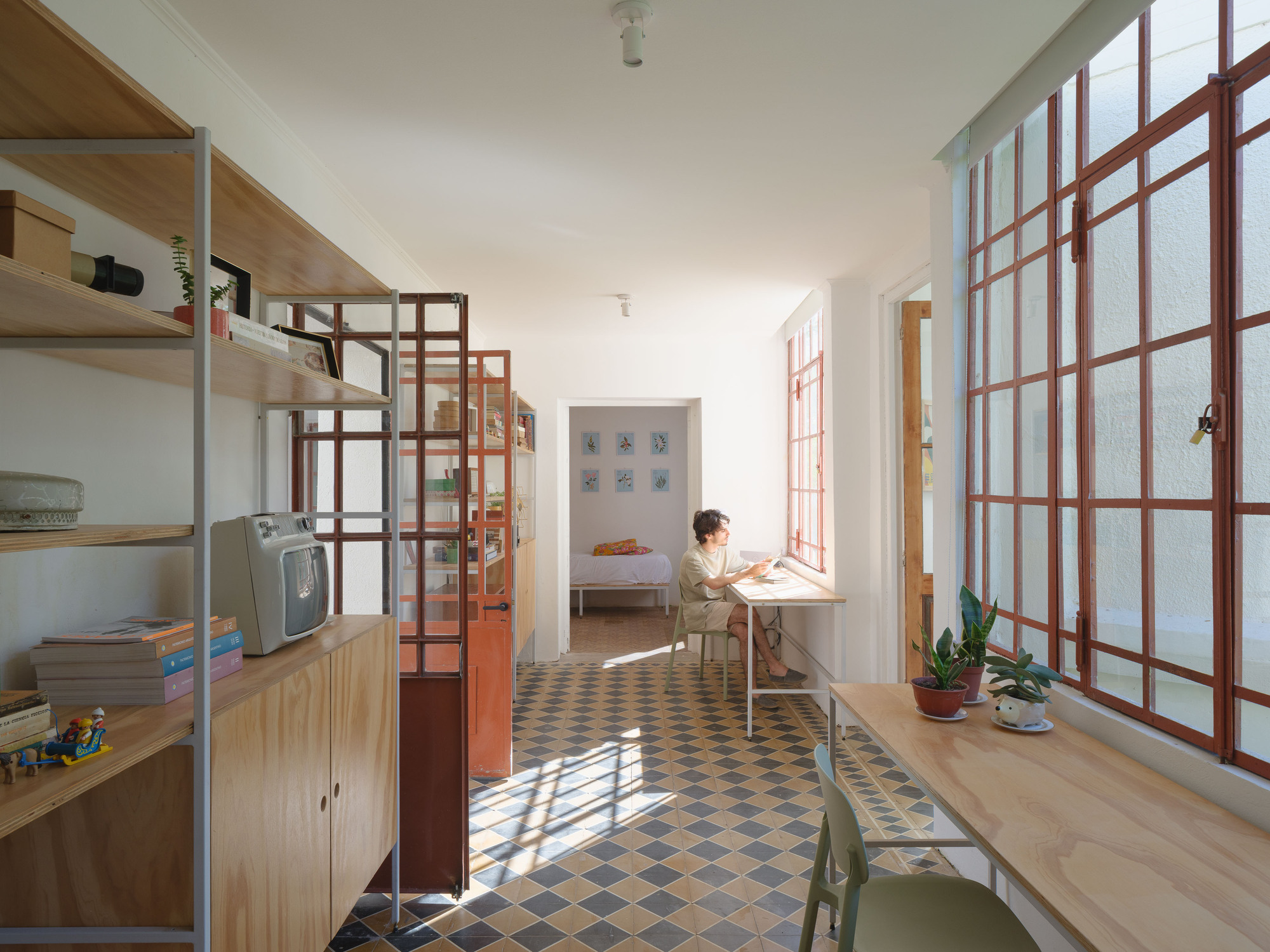 Long hallway with chequered floor tiles, red-framed glass partitions and a person reading at a window desk