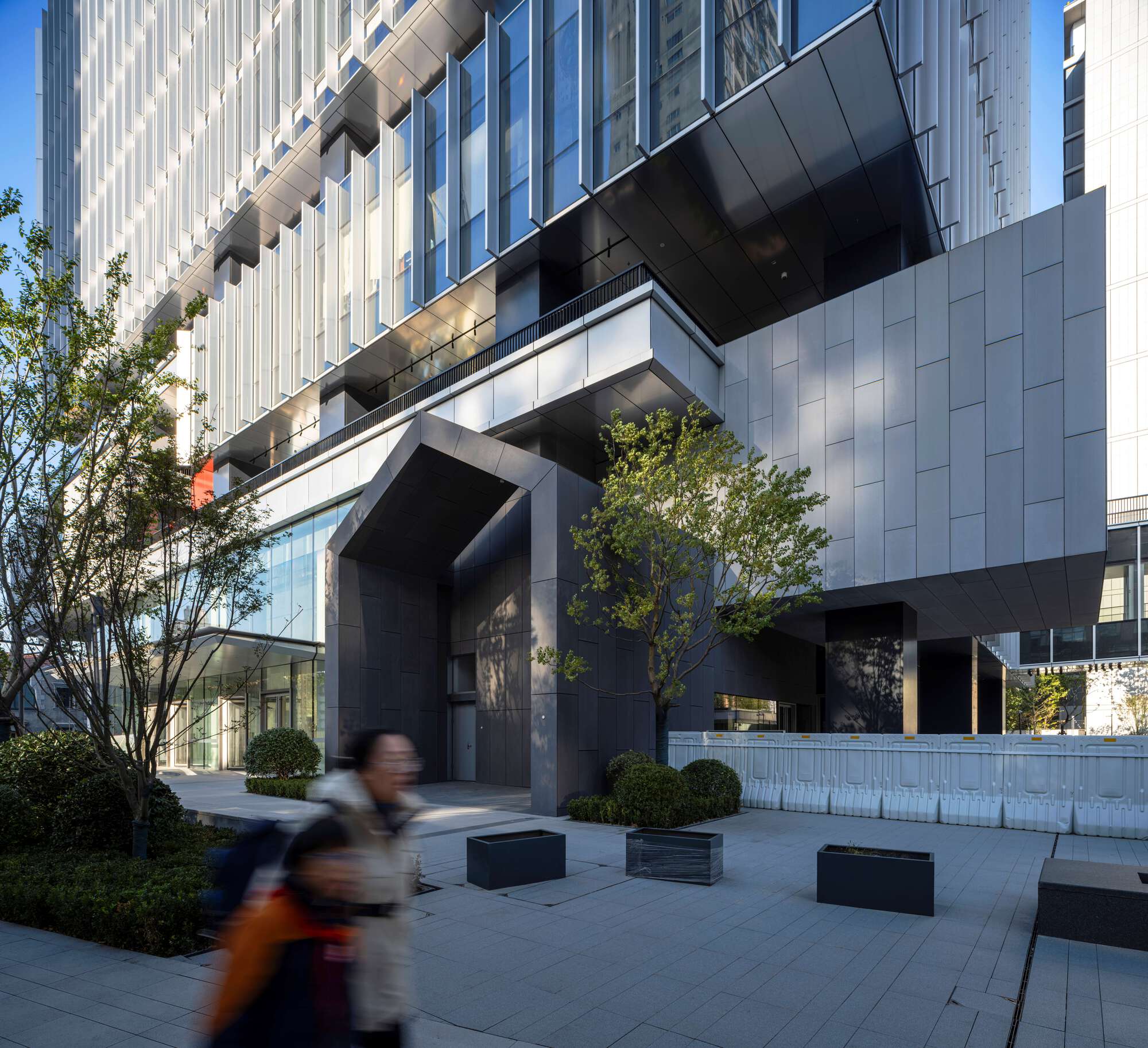 Ground-level plaza with black benches and planted beds as a person walks beneath the cantilevered volume