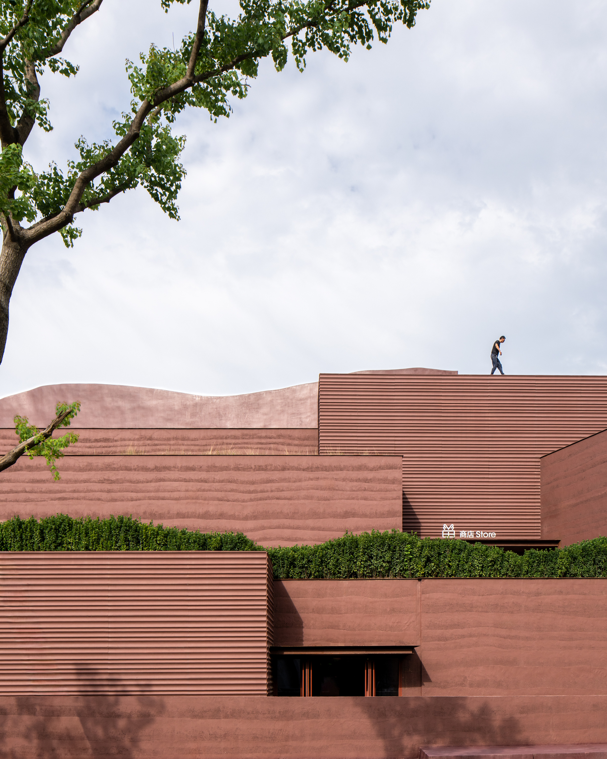 Layered red stone facades with horizontal ribbing and a person walking the roofline under tree branches