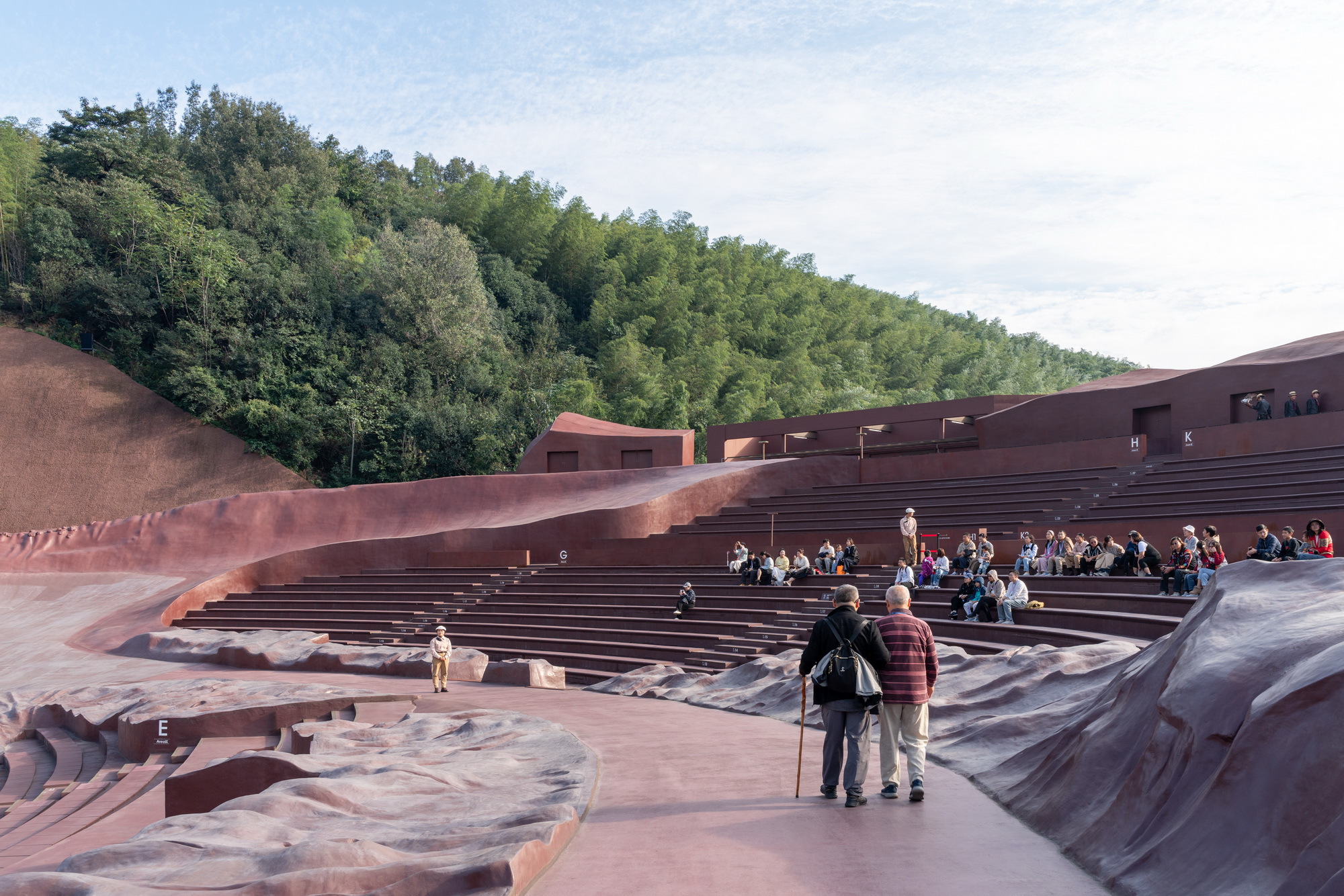 Visitors seated in the amphitheatre bowl with terraced red stone seating facing the wooded slope