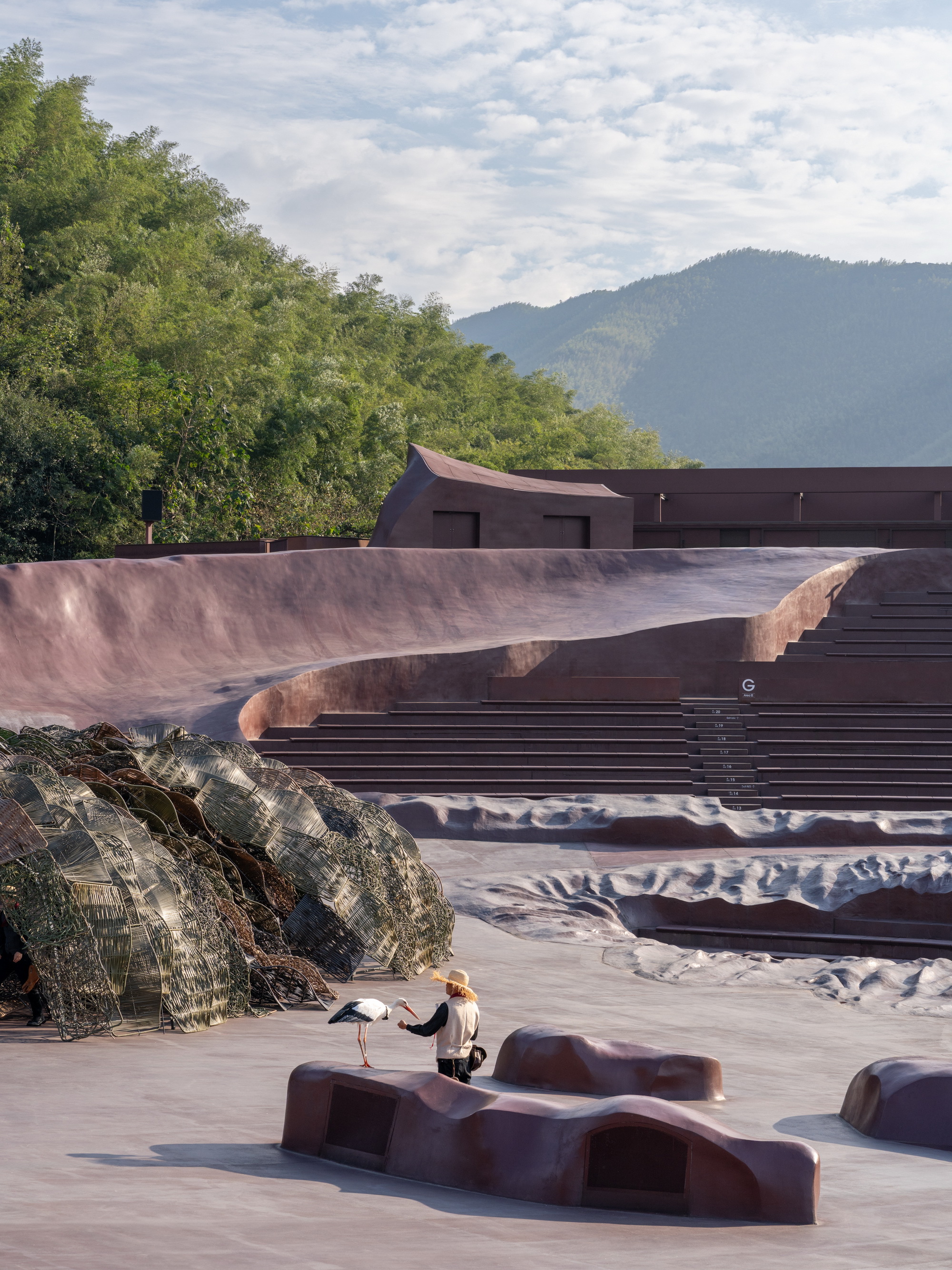 Outdoor courtyard with sculptural seating elements and a visitor beneath cloudy skies and distant peaks