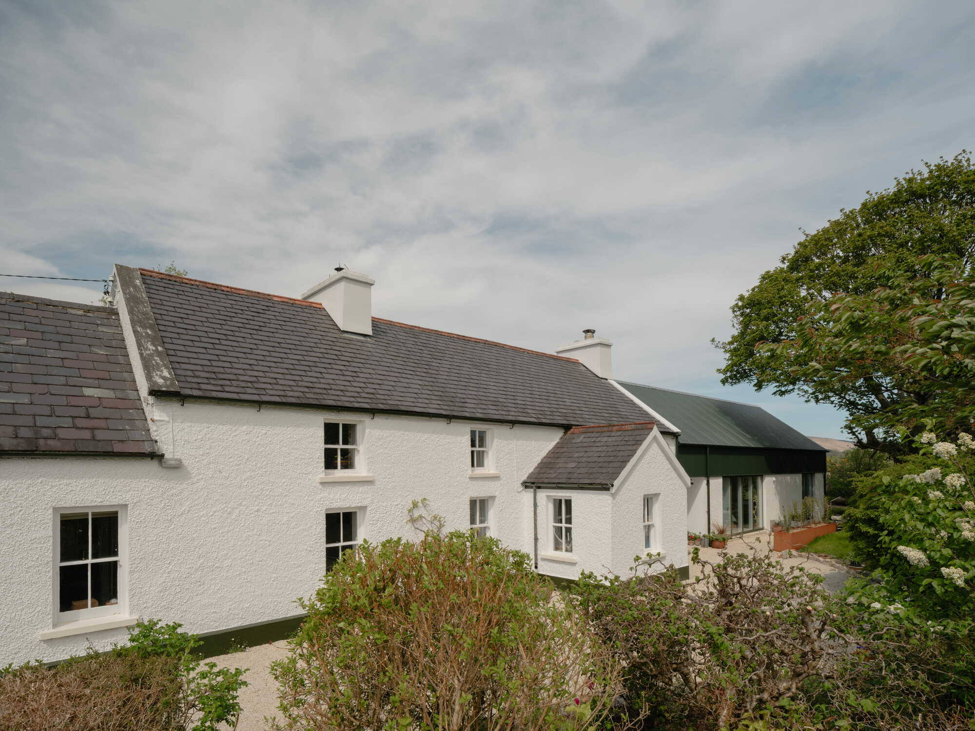 Whitewashed cottage with grey tiled roofs and a green corrugated metal extension under cloudy skies