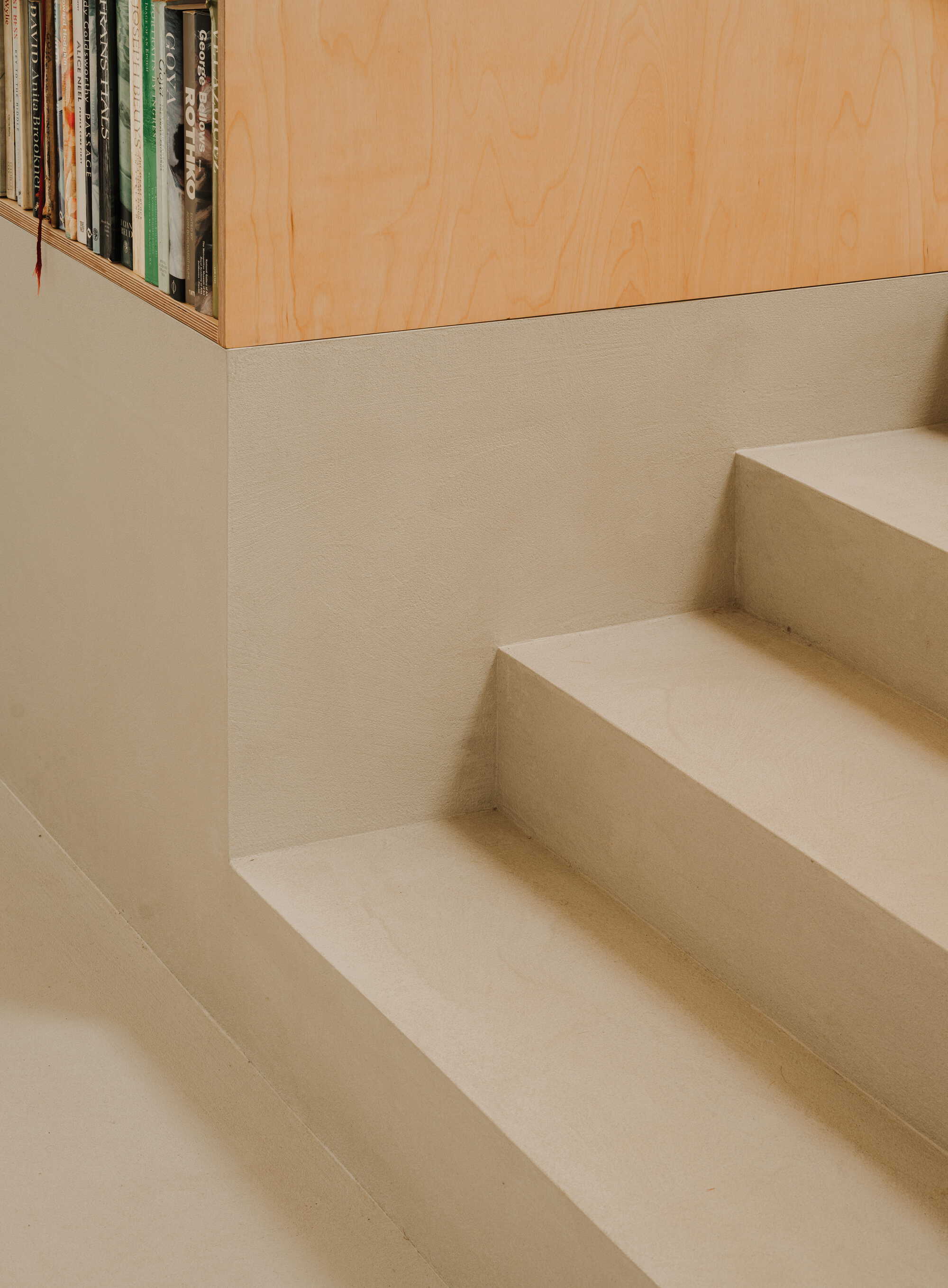 Detail of plaster staircase treads meeting a white wall beneath a plywood bookshelf
