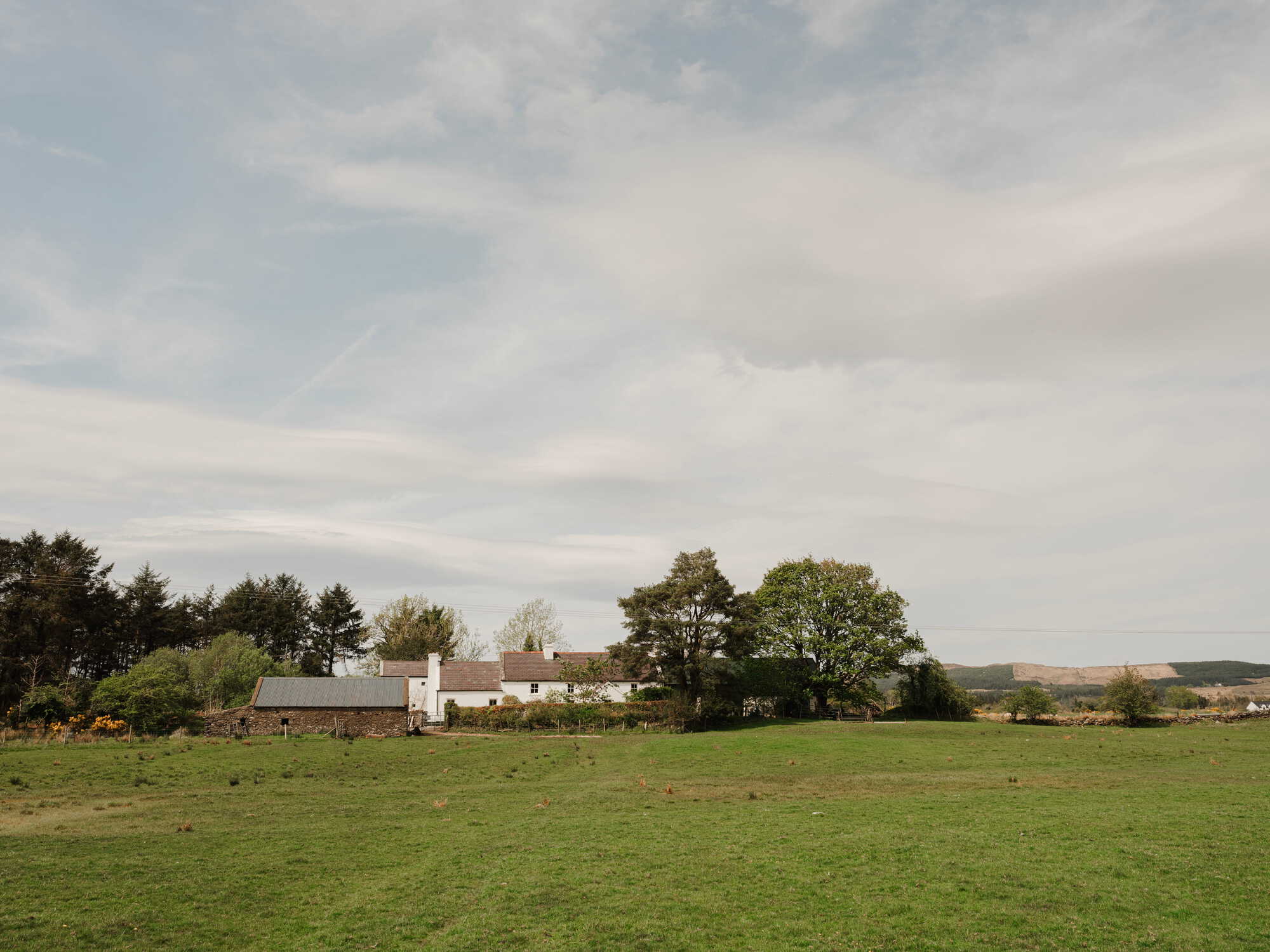 Low whitewashed farmhouse in a grass field with mature trees under an overcast sky