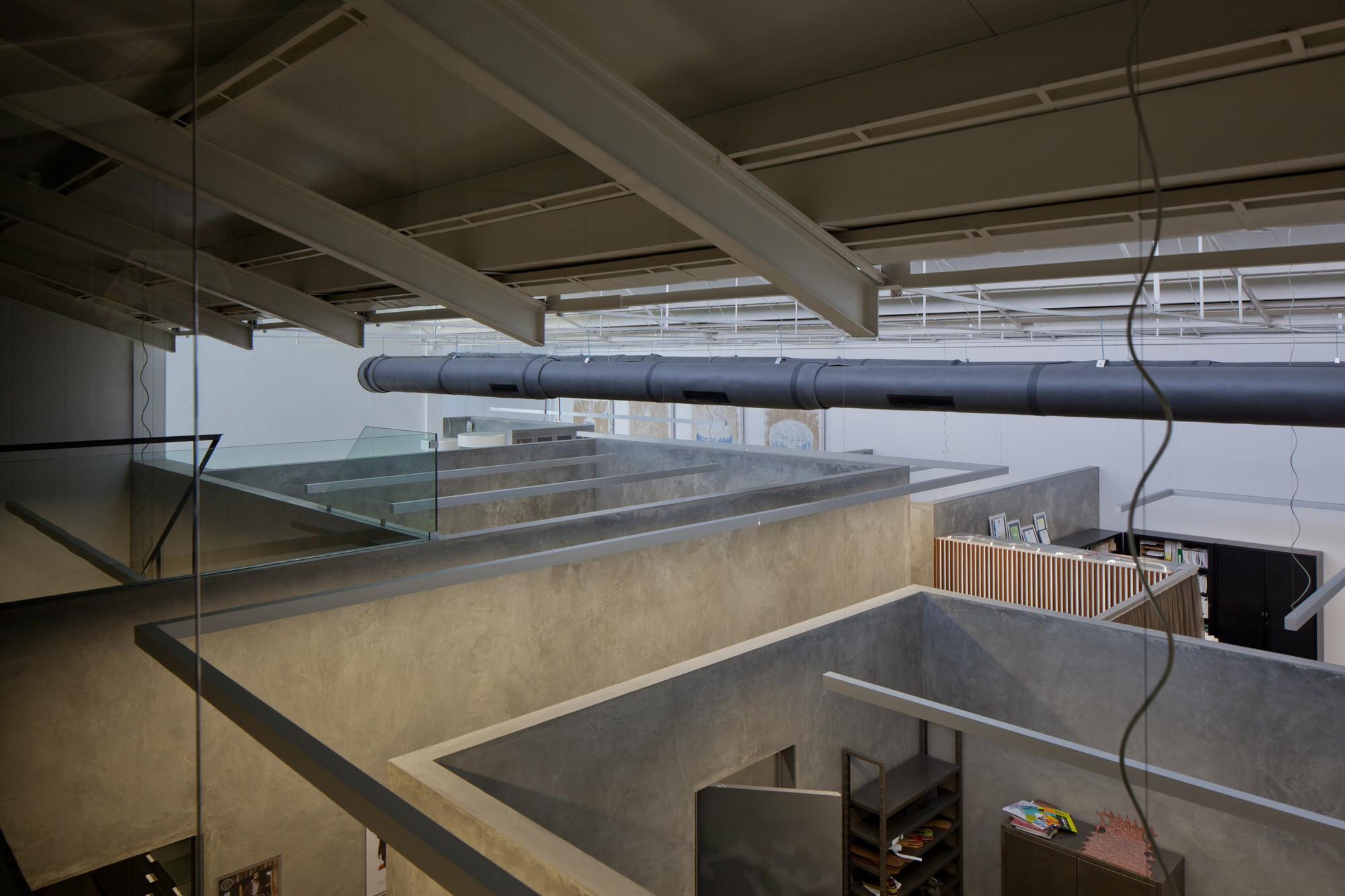 View from the upper level across glass balustrades and plastered walls toward a dark suspended duct and shelving below