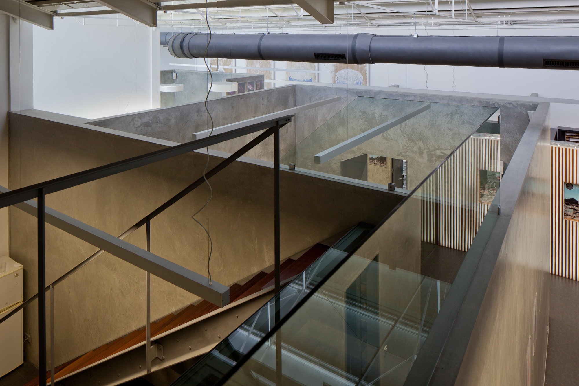 Steel and glass balustrade above a wood-tread staircase, with a grey duct overhead and timber slat walls beyond