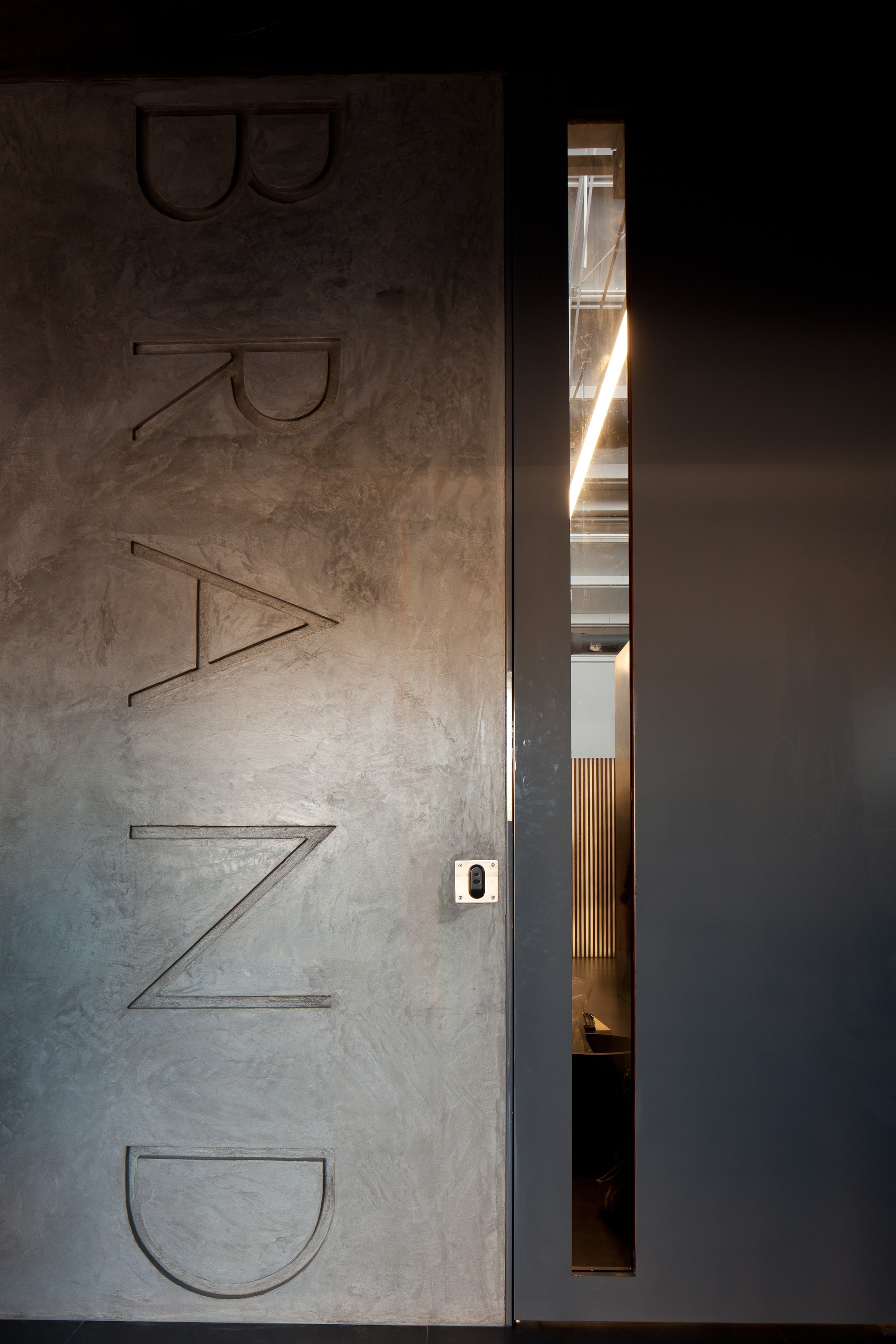 Plastered wall with vertical embossed BRAND lettering beside a dark door with a narrow glass slot showing a lit corridor