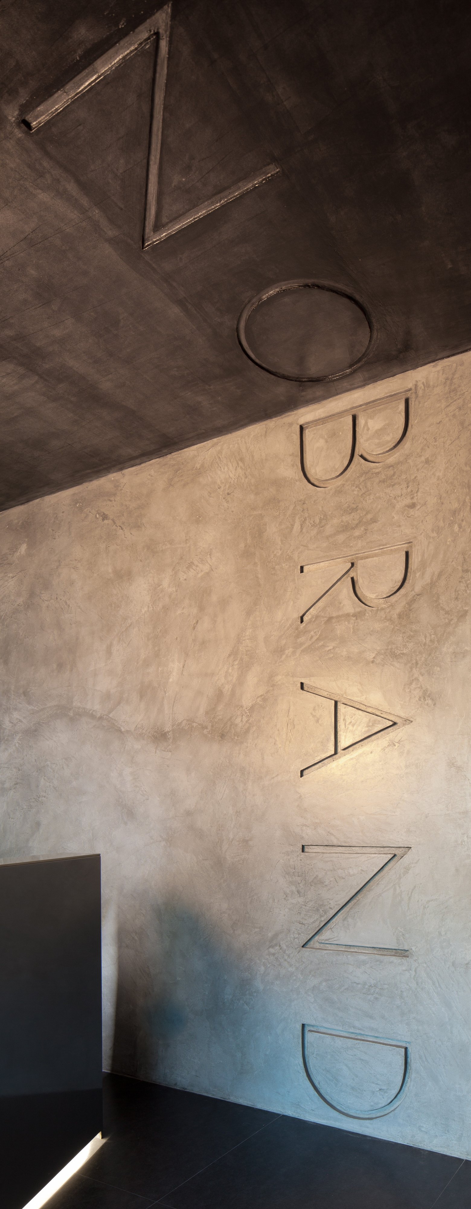 Embossed NO BRAND letters running from the textured plaster wall onto the dark ceiling, with a strip of light at the floor