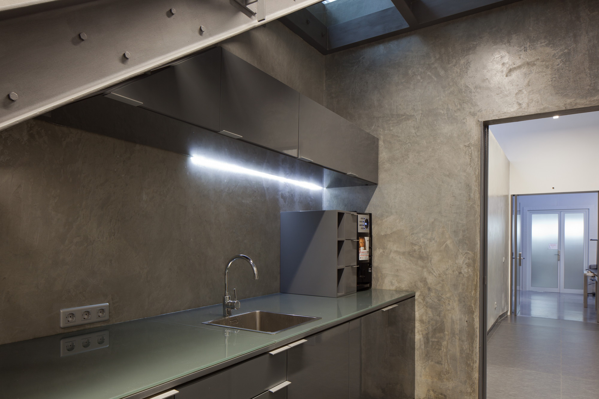 Small kitchenette with a steel sink, grey cabinets and an under-cabinet light strip against plaster walls, a doorway opening to a corridor