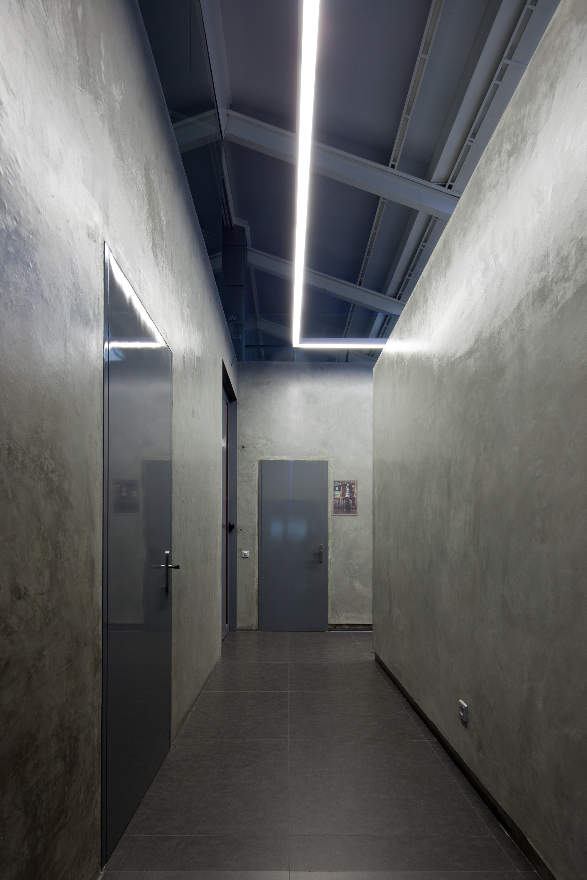 Narrow corridor with rough plastered concrete-look walls, grey doors and a long linear light strip running along exposed steel roof trusses