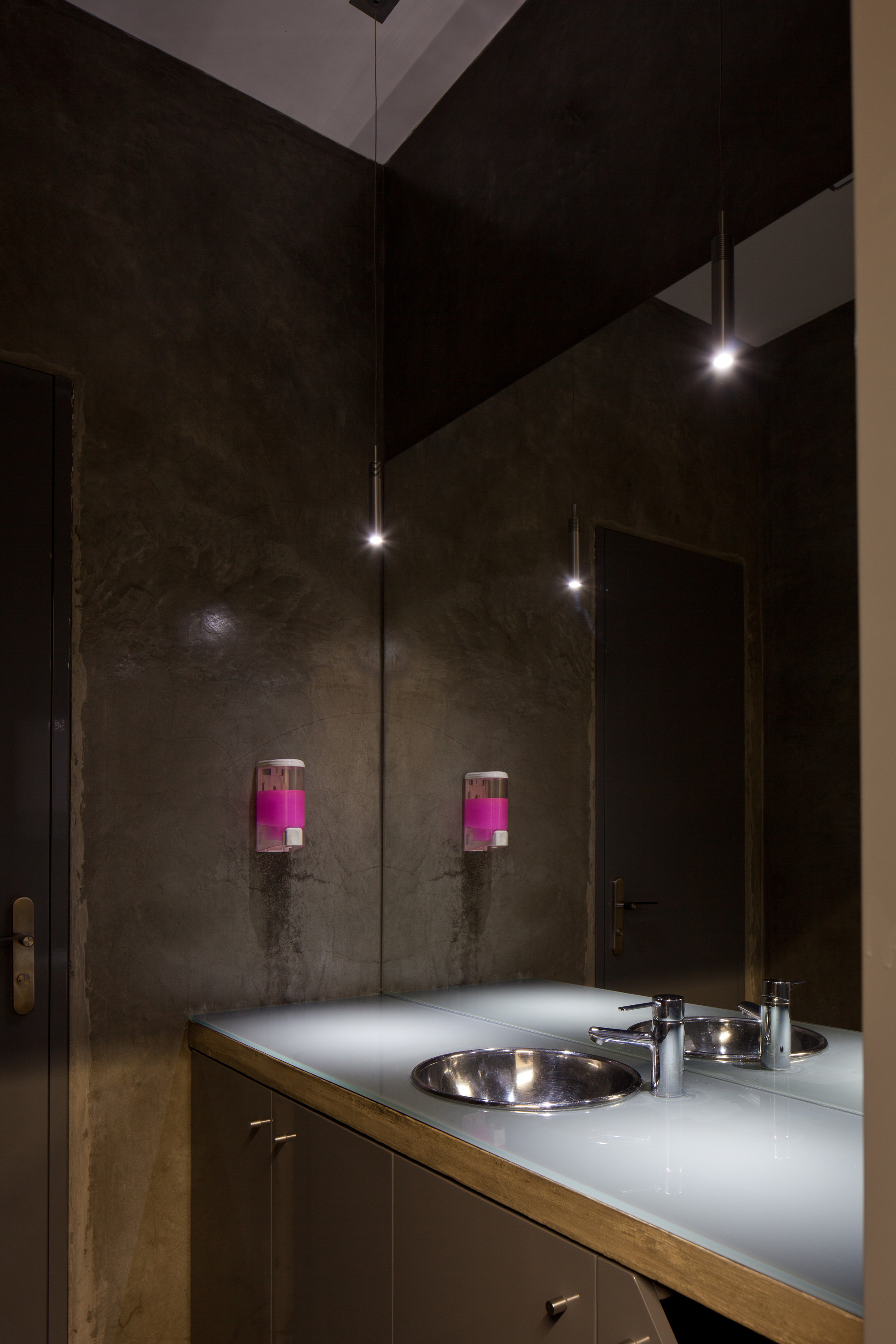 Washroom with a glass-topped vanity, steel basin, pink soap dispensers and pendant lights reflected in a full-height mirror on dark plaster walls