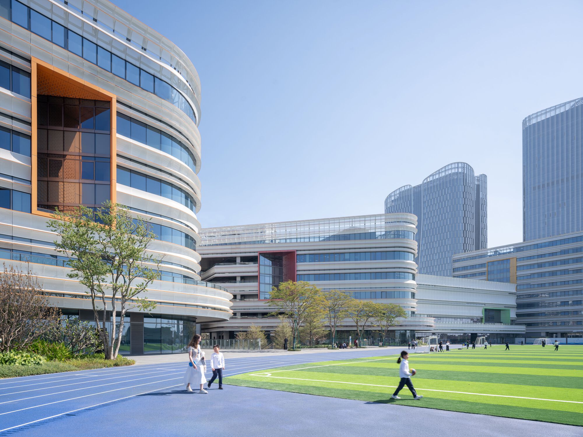 Perforated orange panel framing the entrance with pedestrians crossing the running track in afternoon light