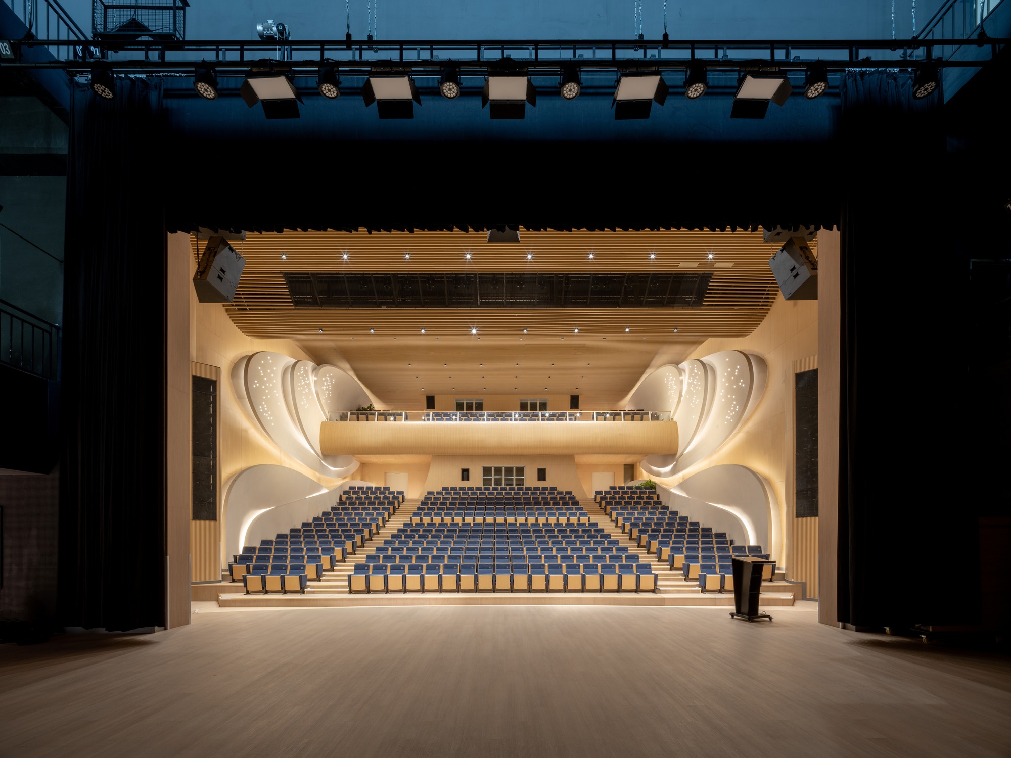 View from the stage into the auditorium showing curved balconies, timber acoustic ceiling, and rows of blue seats