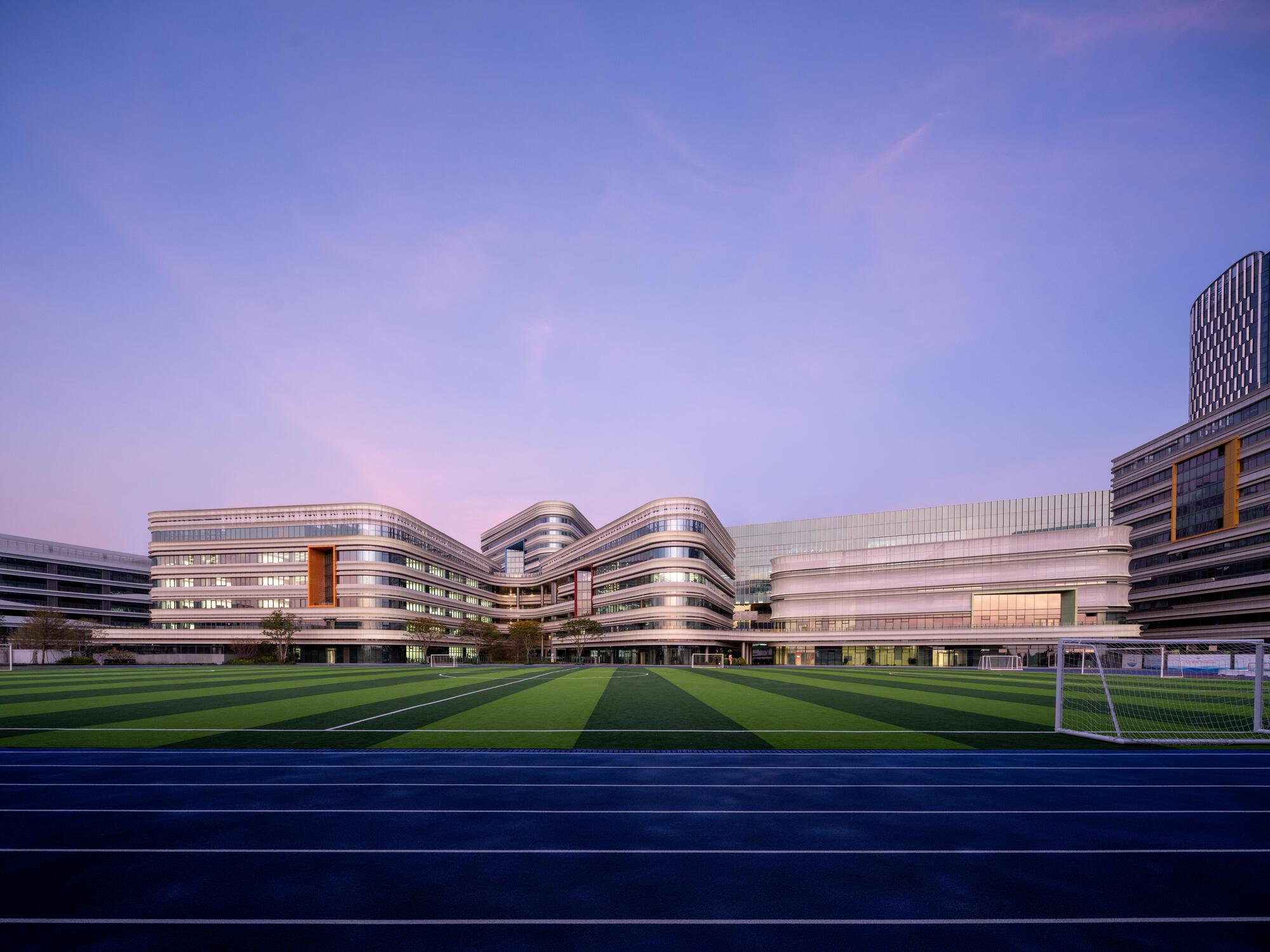 View across the sports pitch toward the banded facades and curved volumes at dusk