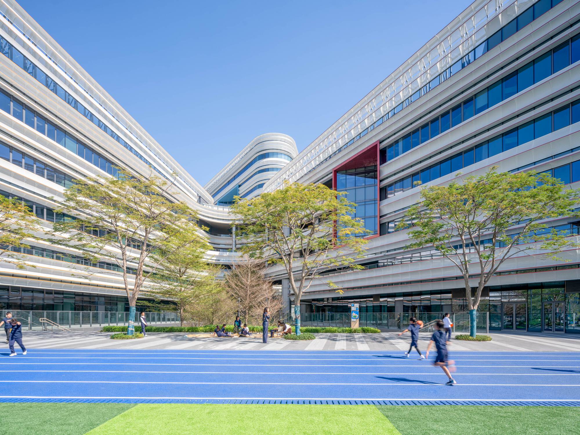 Courtyard with blue running track and people walking among trees framed by horizontally striped glass facade volumes