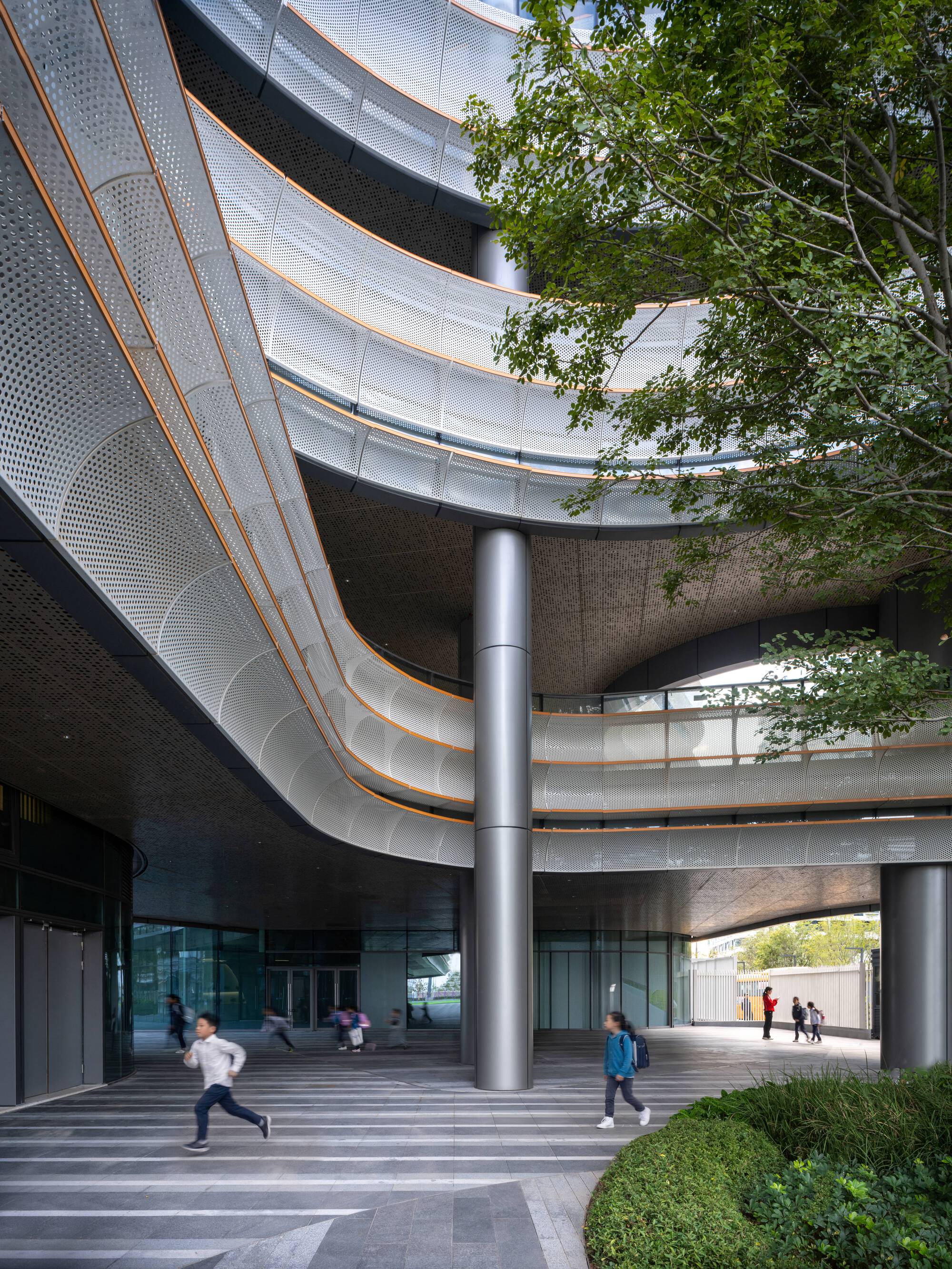 Covered entry plaza with sweeping perforated metal canopy, cylindrical columns, and children running past planted beds