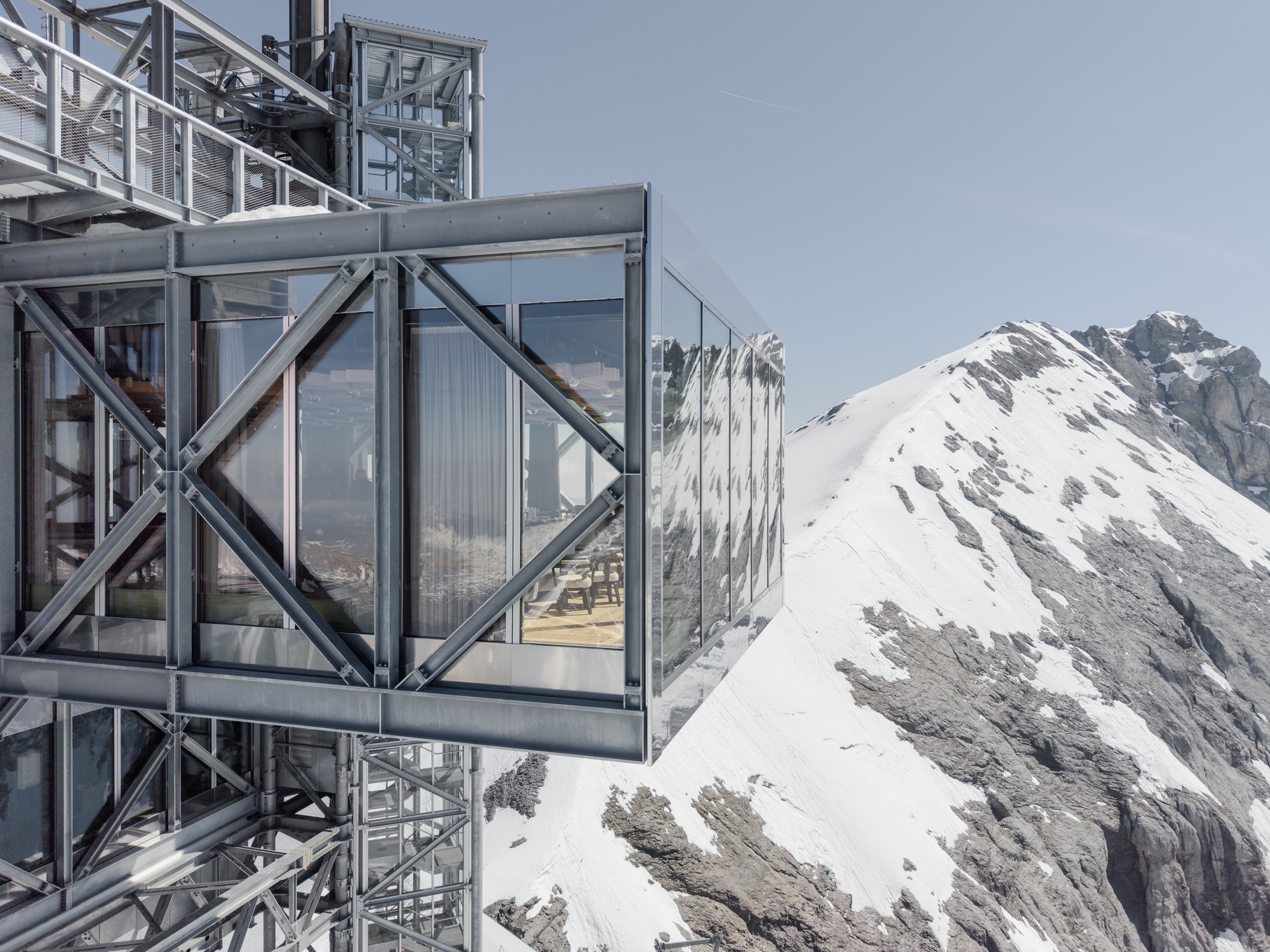 Glass-enclosed viewing platform cantilevered from the steel frame with mountain peaks beyond