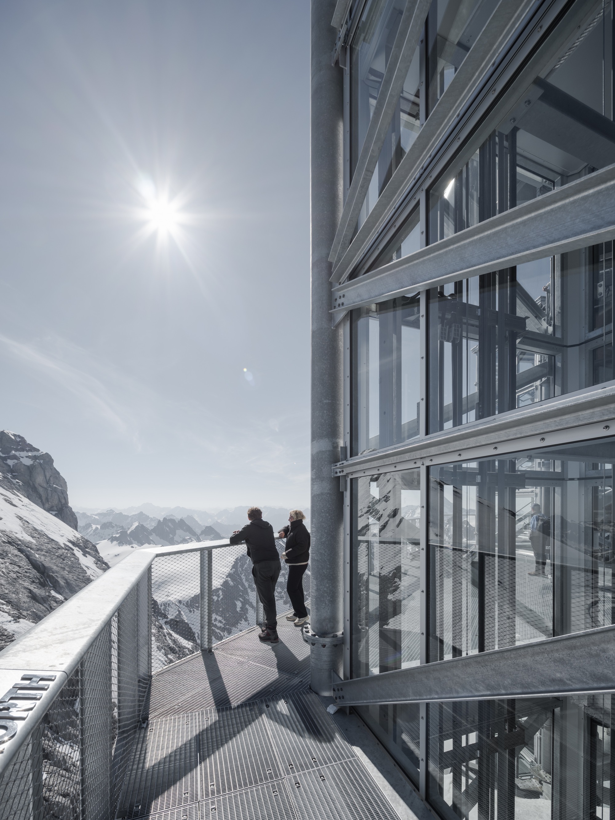 Two visitors on the exterior metal walkway beside the glazed tower under bright sun