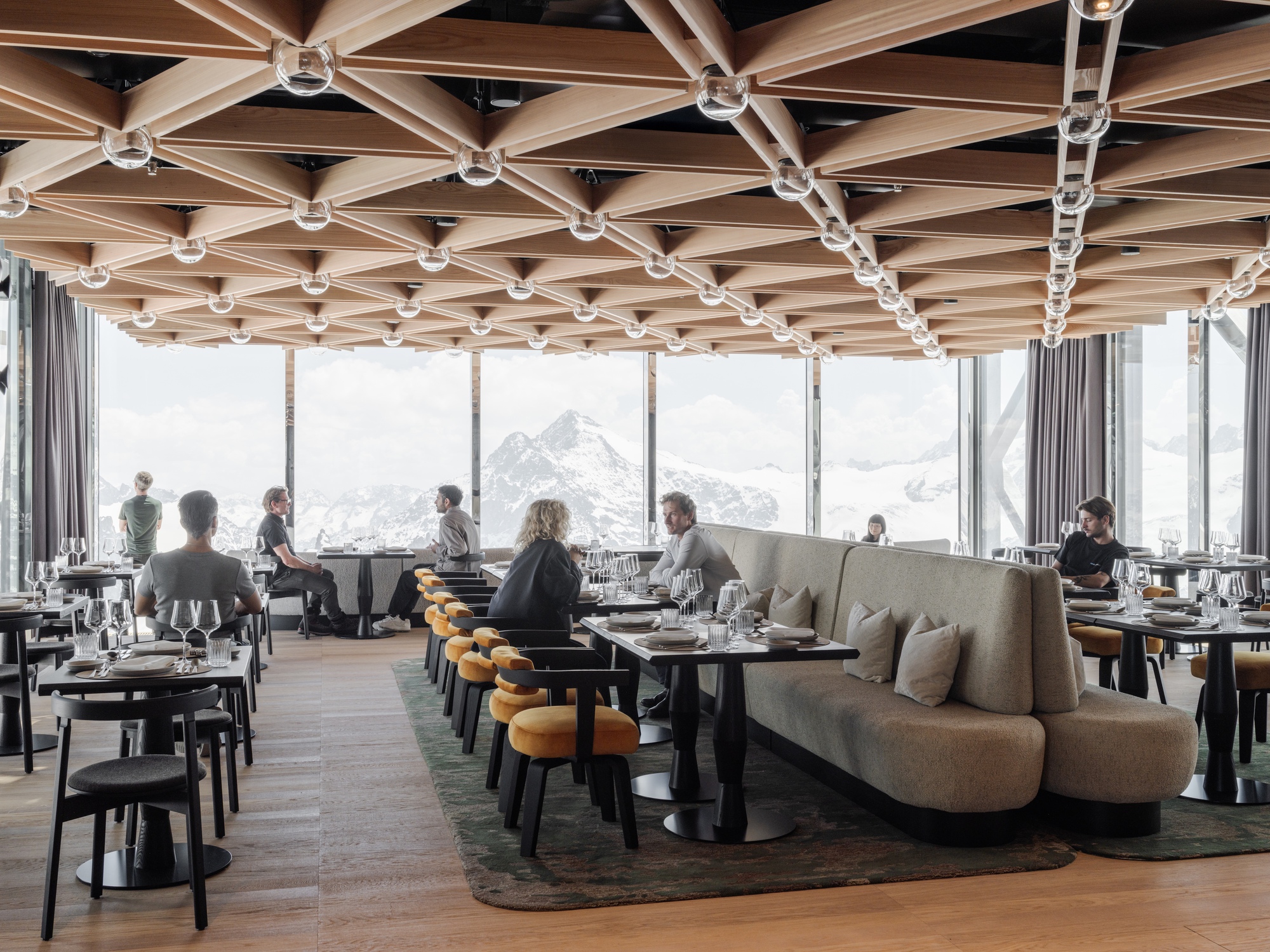 Restaurant interior with geometric timber ceiling grid and floor-to-ceiling windows overlooking snow peaks