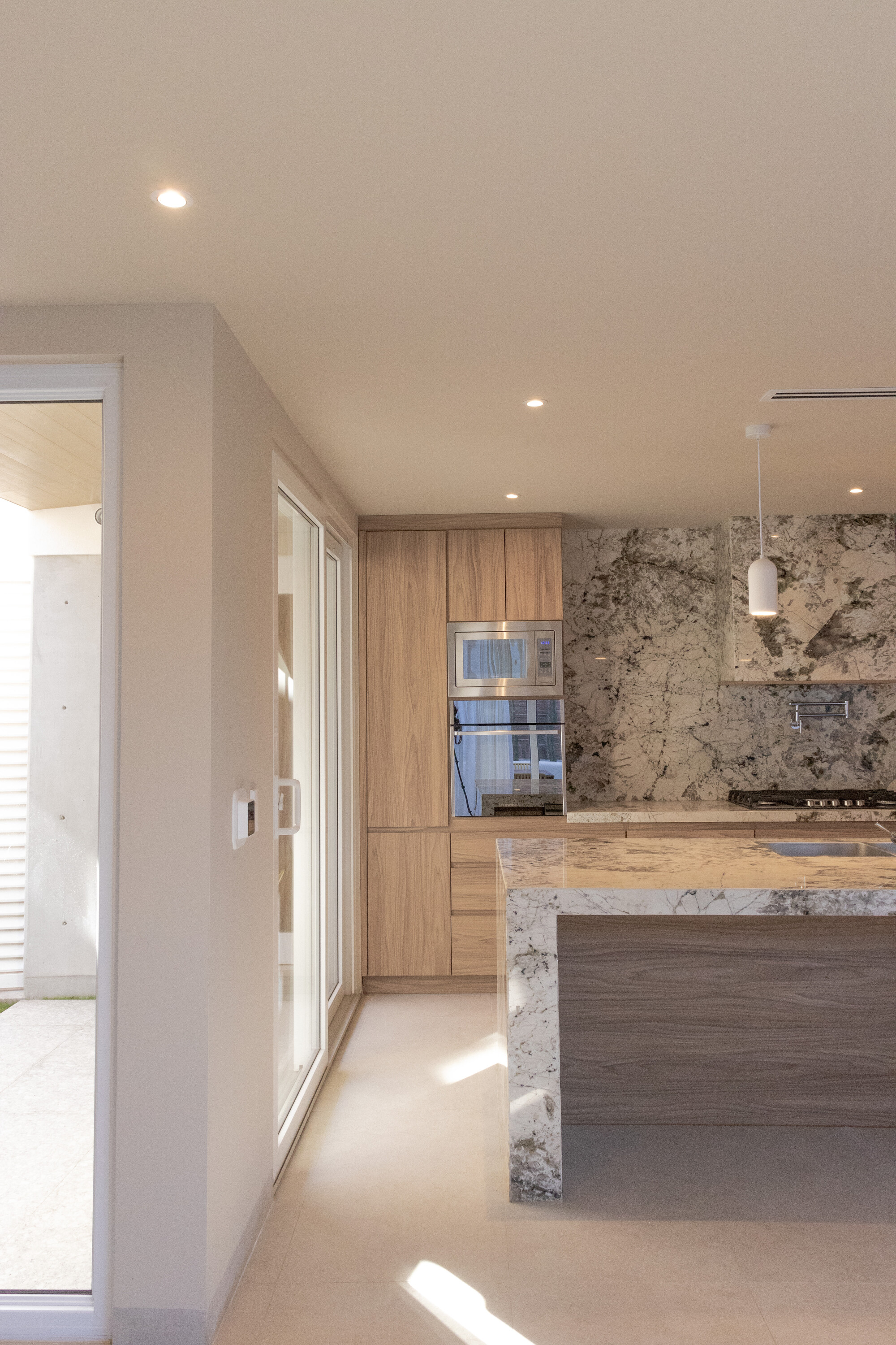 Kitchen with pale stone countertop, timber cabinetry and recessed lighting through glazed doors