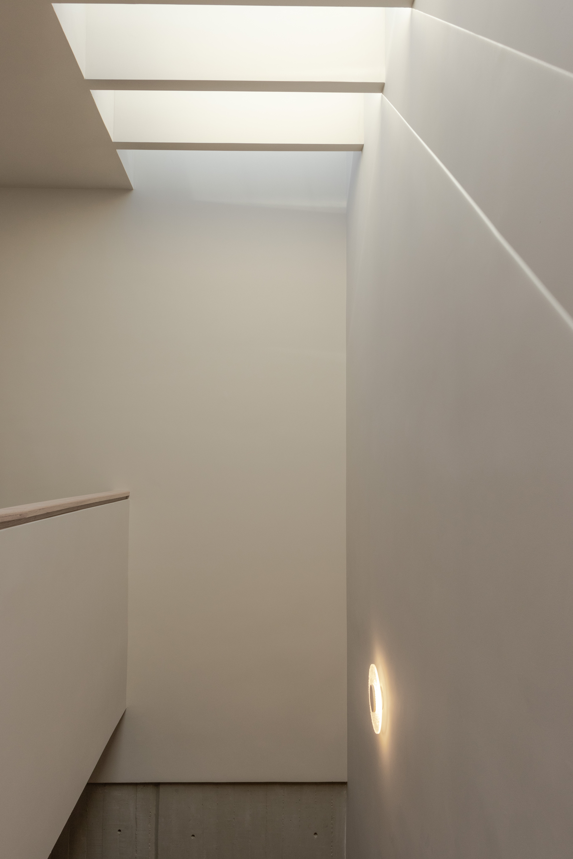 Stairwell with slotted skylight casting linear shadows across white walls and concrete floor