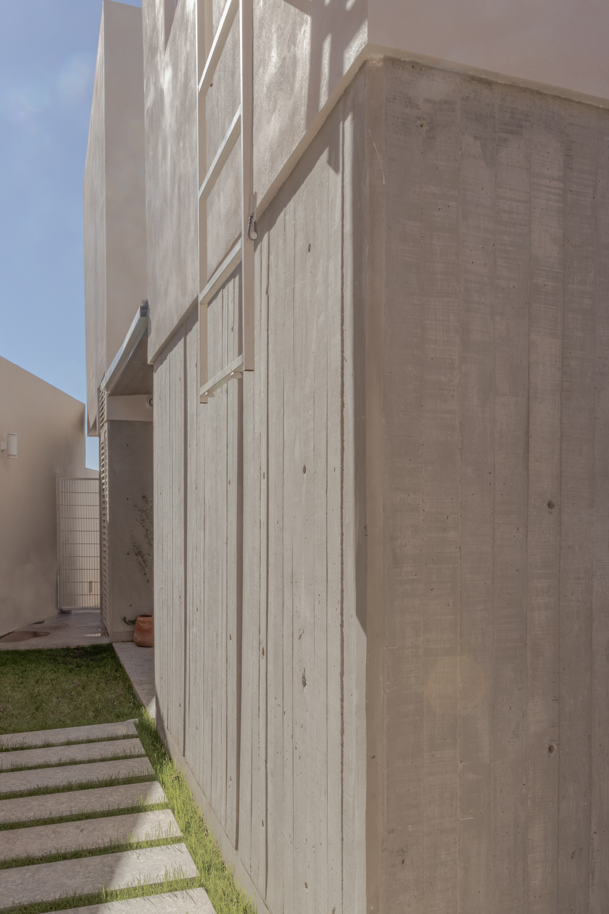 Boardmarked concrete facade with projecting horizontal louvers beside a grass pathway in sunlight