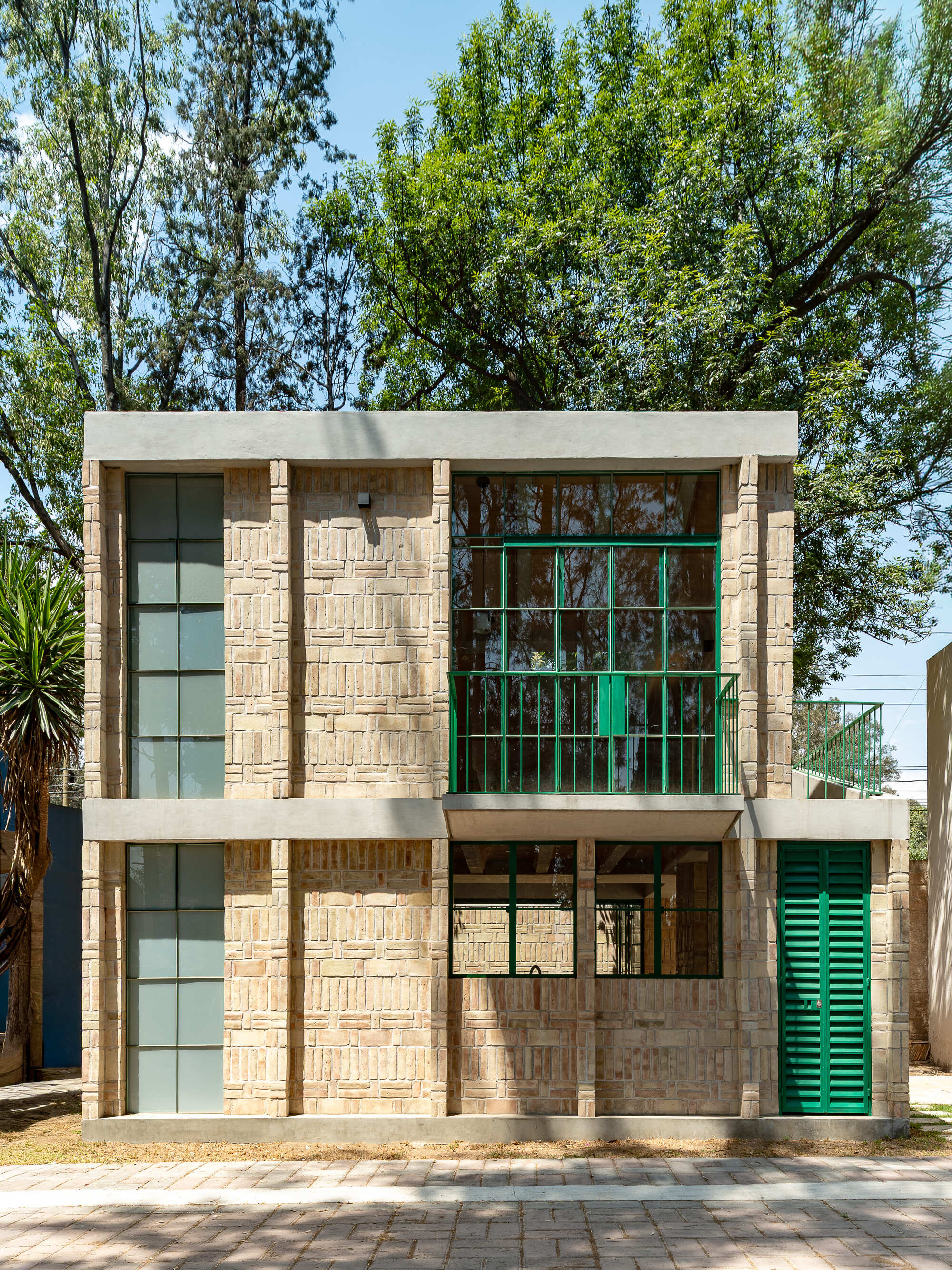 Street facade with textured brick walls, green metal-framed windows, and concrete bands framed by tall trees