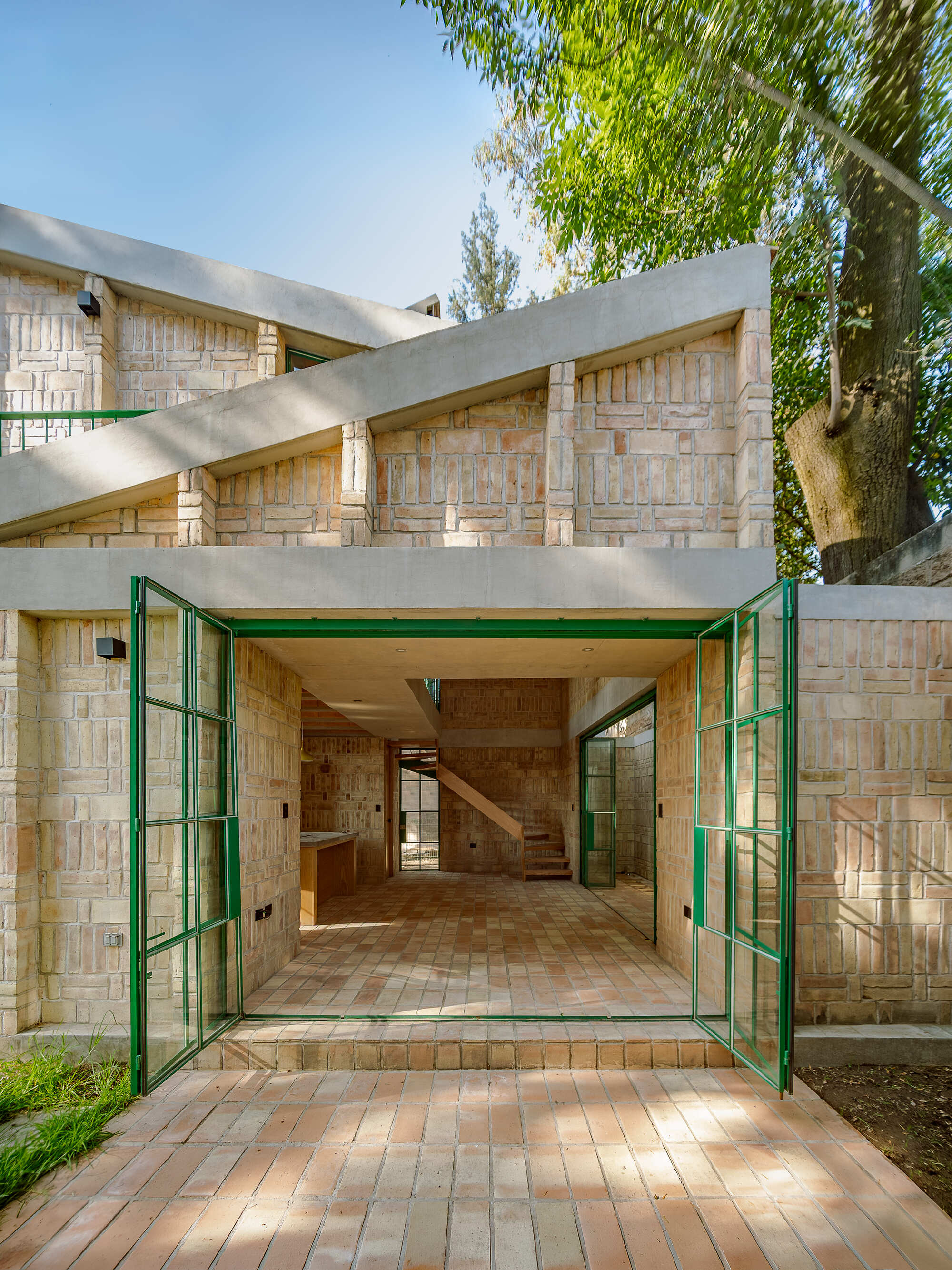 Open green metal-framed doors revealing the brick-paved interior and staircase beyond under dappled tree shade