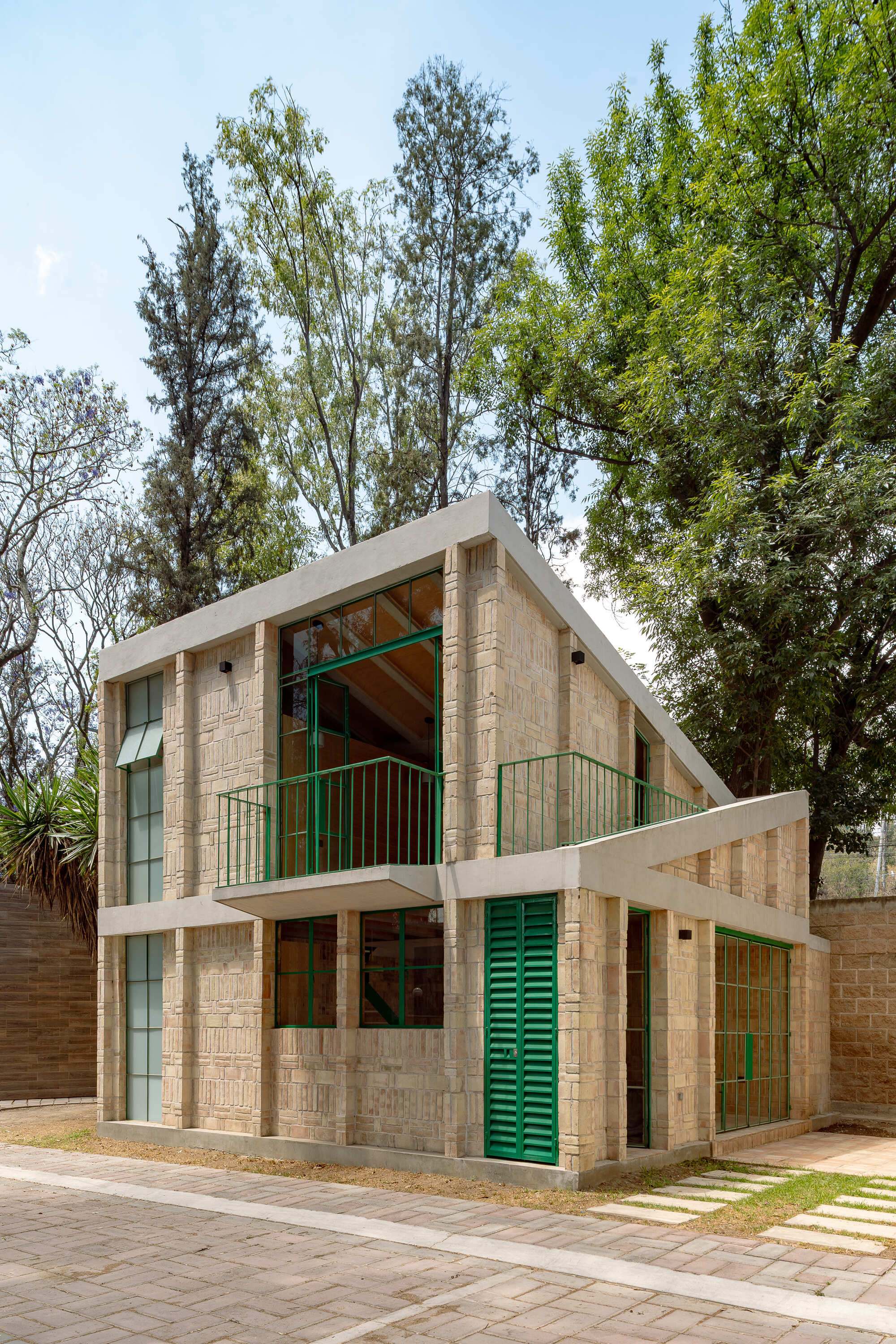 Street view of the two-story textured brick house with green shutters and balcony among tall trees
