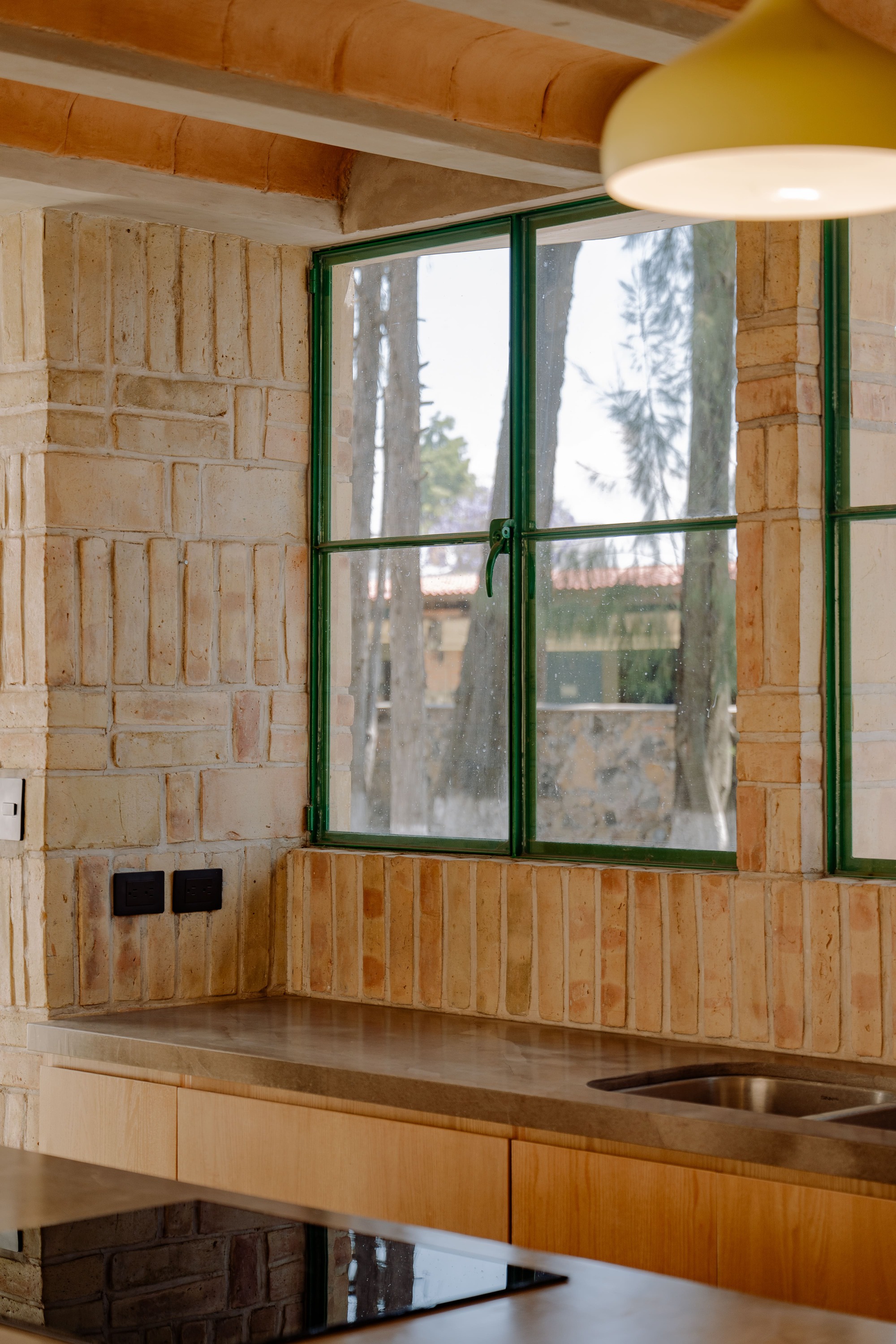 Kitchen counter with green-framed window in textured brick wall under timber ceiling and pendant light