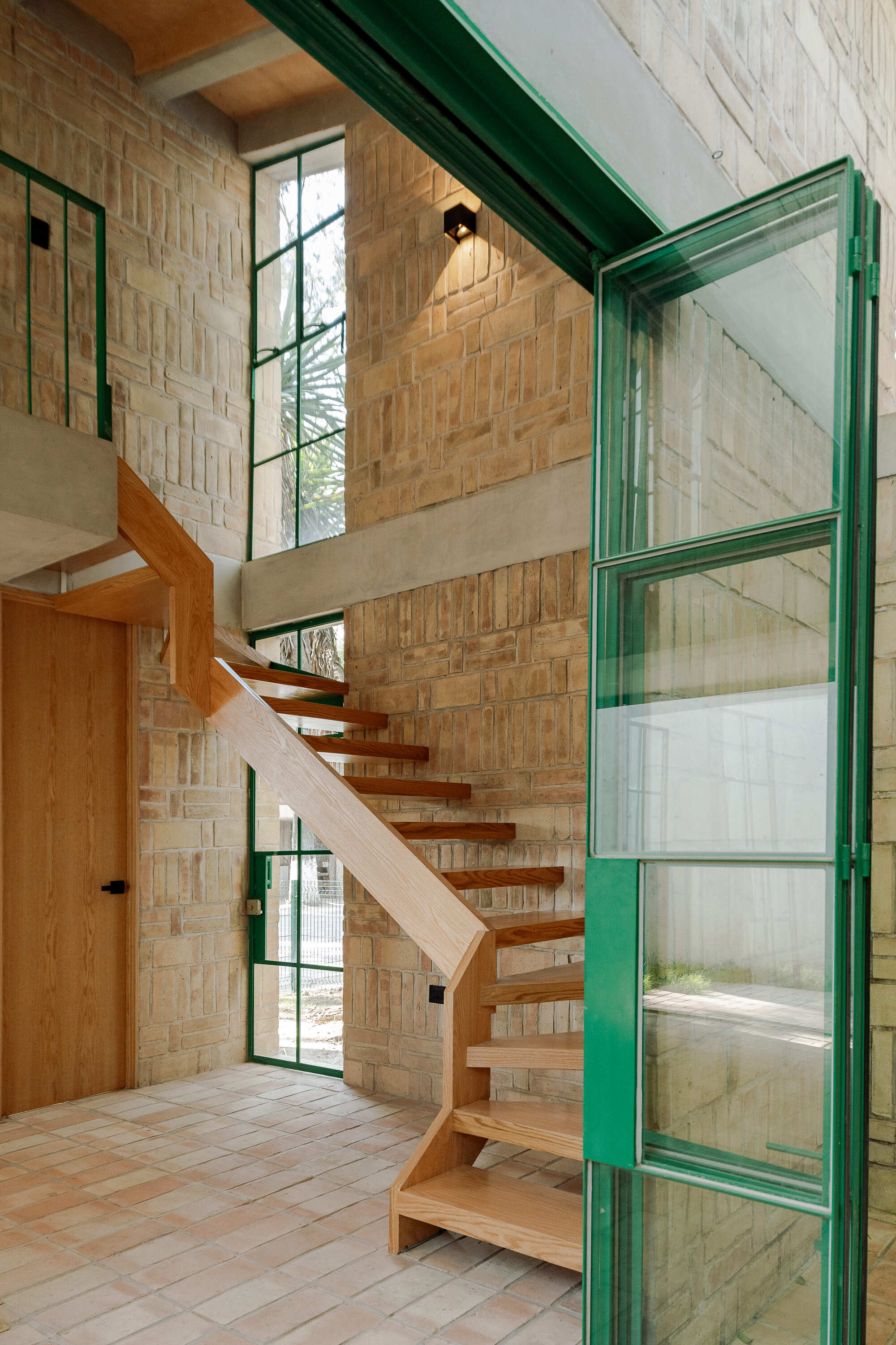 Double-height stairwell with timber treads, green steel door and pale brick walls in soft daylight