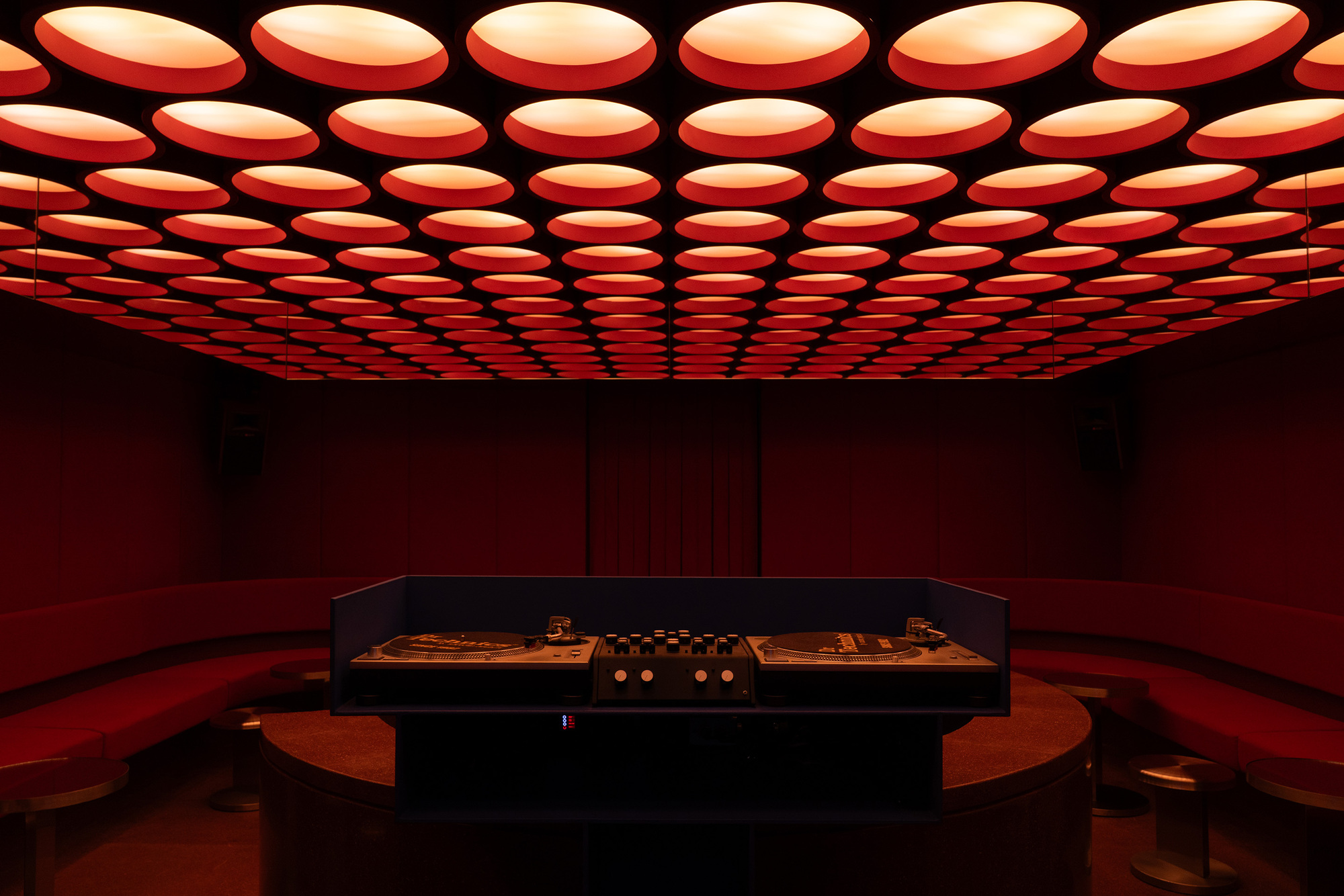 Central display counter with circular orange ottomans beneath the illuminated ceiling grid in the red interior