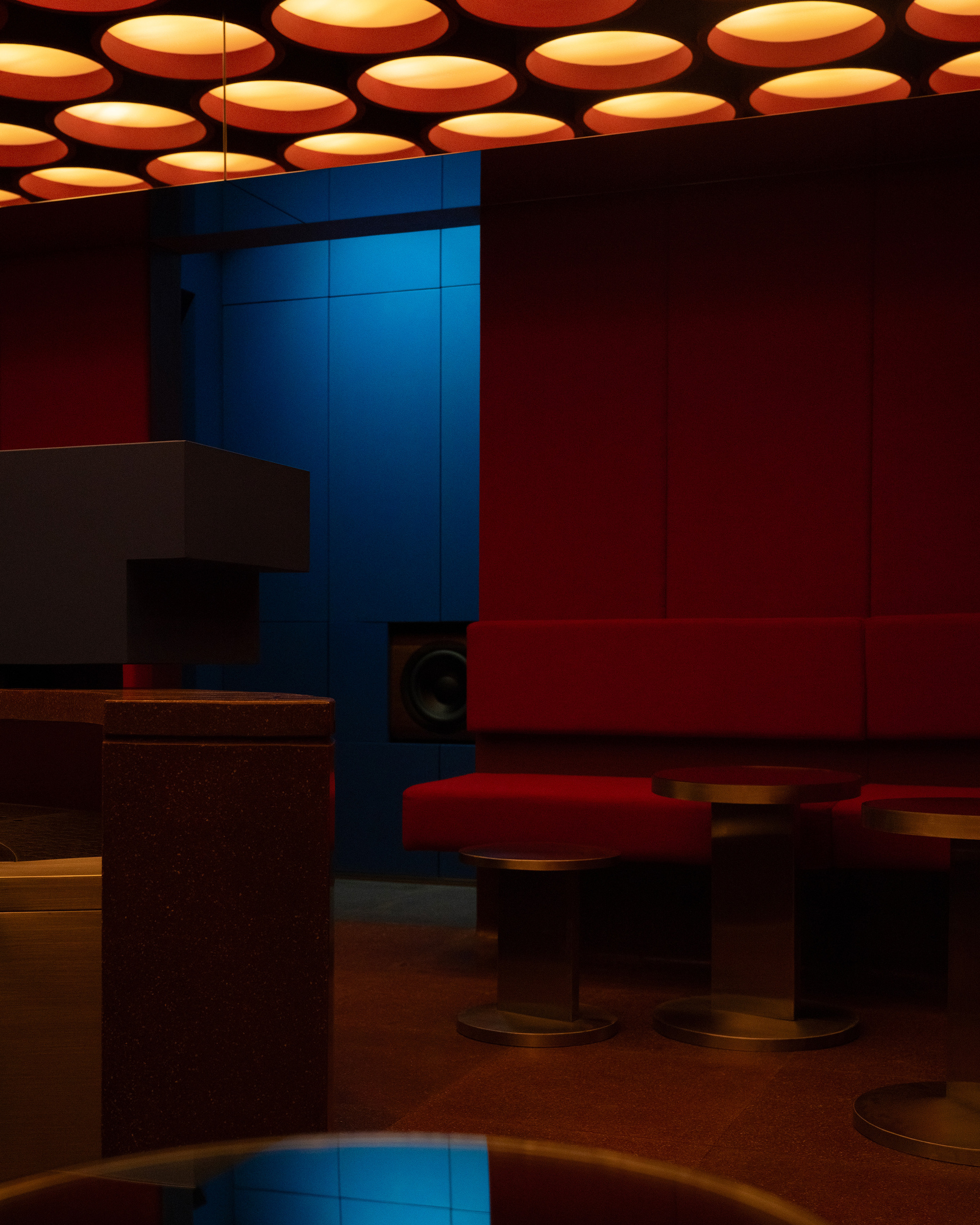 Blue column with built-in speaker beside bronze ottomans and red upholstered seating in low light