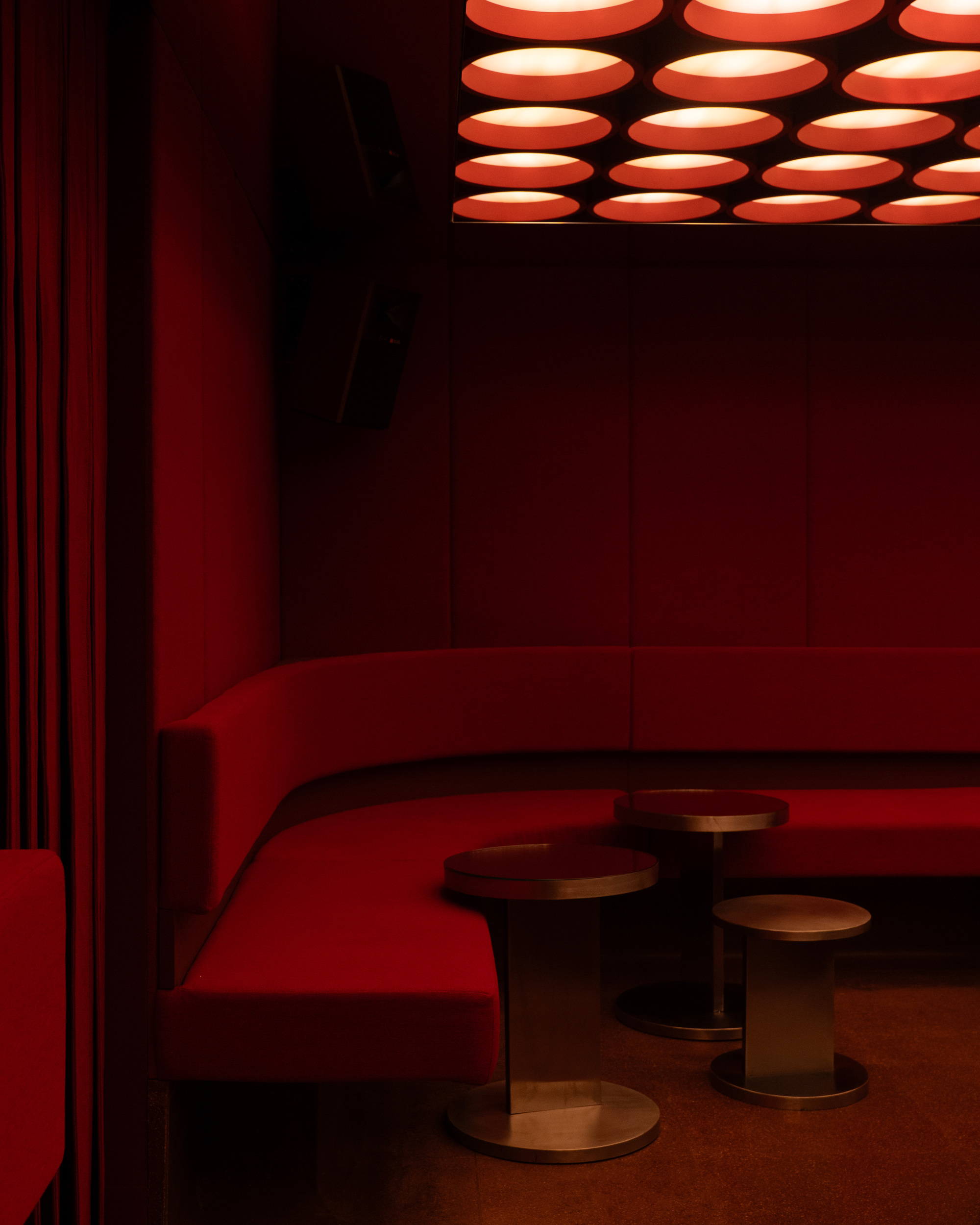 Red pleated curtain beside curved banquette seating with bronze stools under the glowing ceiling grid