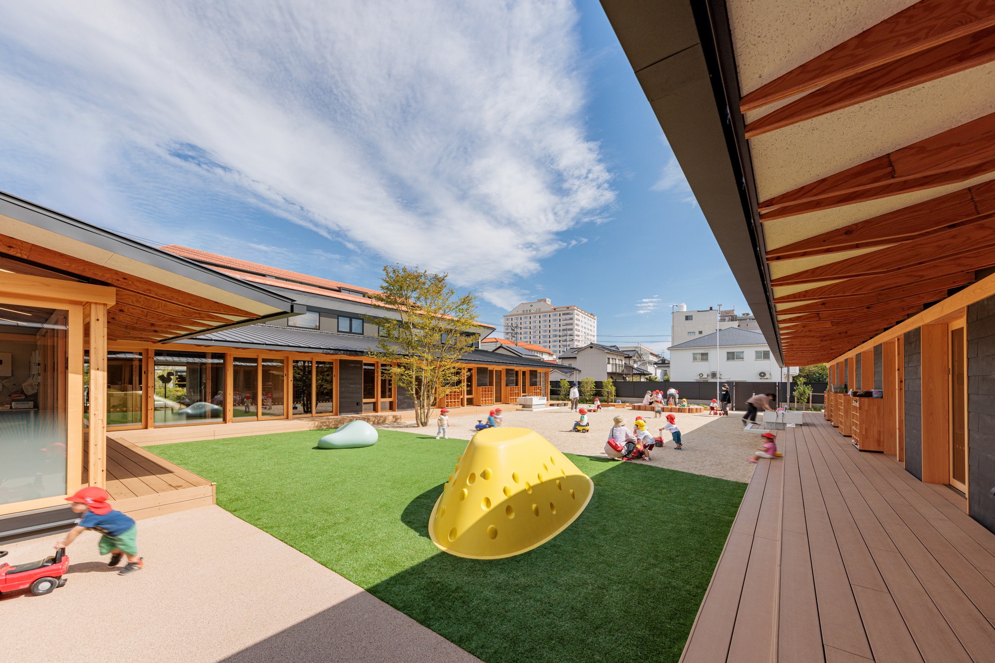 Central courtyard with synthetic turf and play sculptures framed by timber-clad pavilions under blue sky