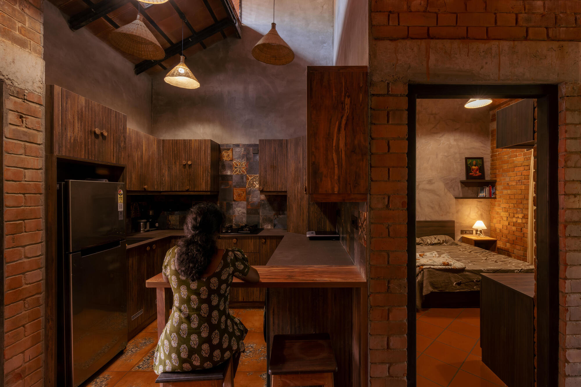 Kitchen with timber cabinetry and terracotta tile wall as a woman prepares food under pendant lights