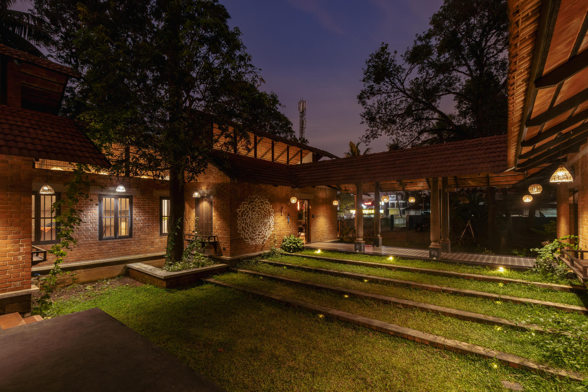 Central courtyard with grass strips and raised planters between brick volumes illuminated by wall lights at twilight