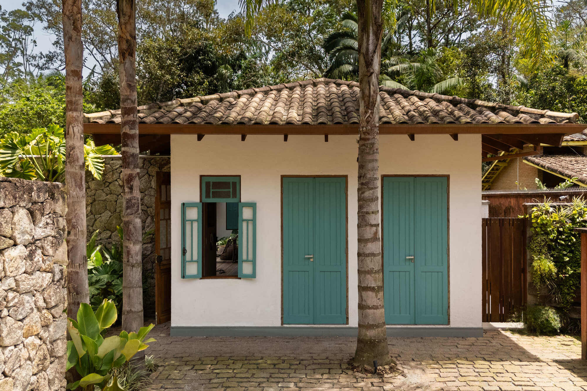 White stucco facade with teal shuttered doors and clay tile roof framed by palms and stone wall