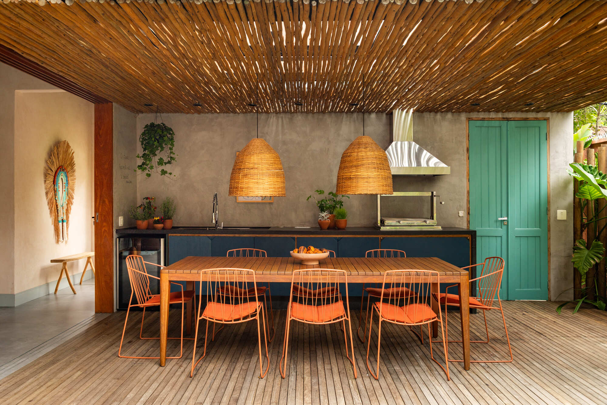 Outdoor kitchen with slatted timber ceiling, woven pendant lamps and turquoise door under overcast sky
