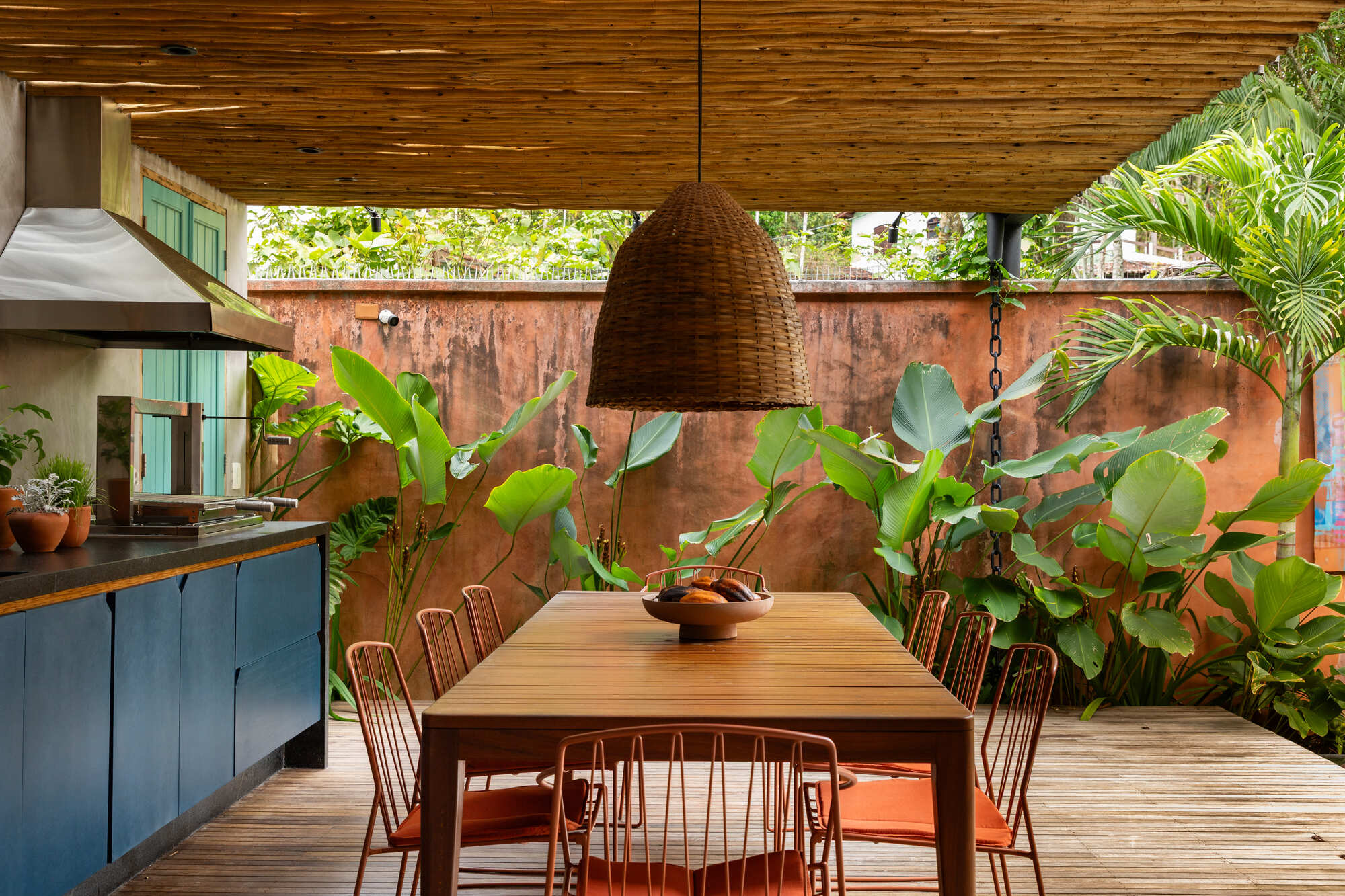 Dining table under woven pendant with weathered copper wall backdrop and banana plants in dappled sunlight