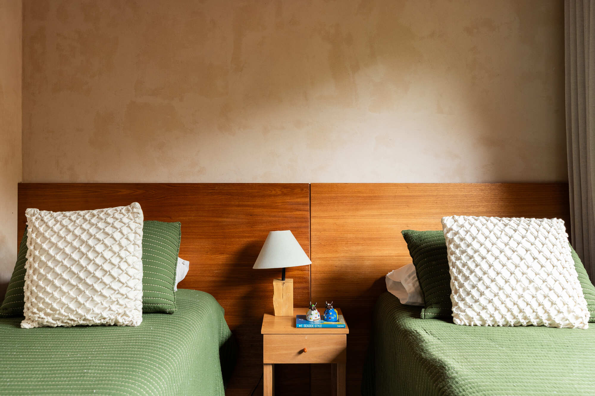Twin beds with timber headboard and central nightstand beneath wall-mounted reading lamp in soft daylight
