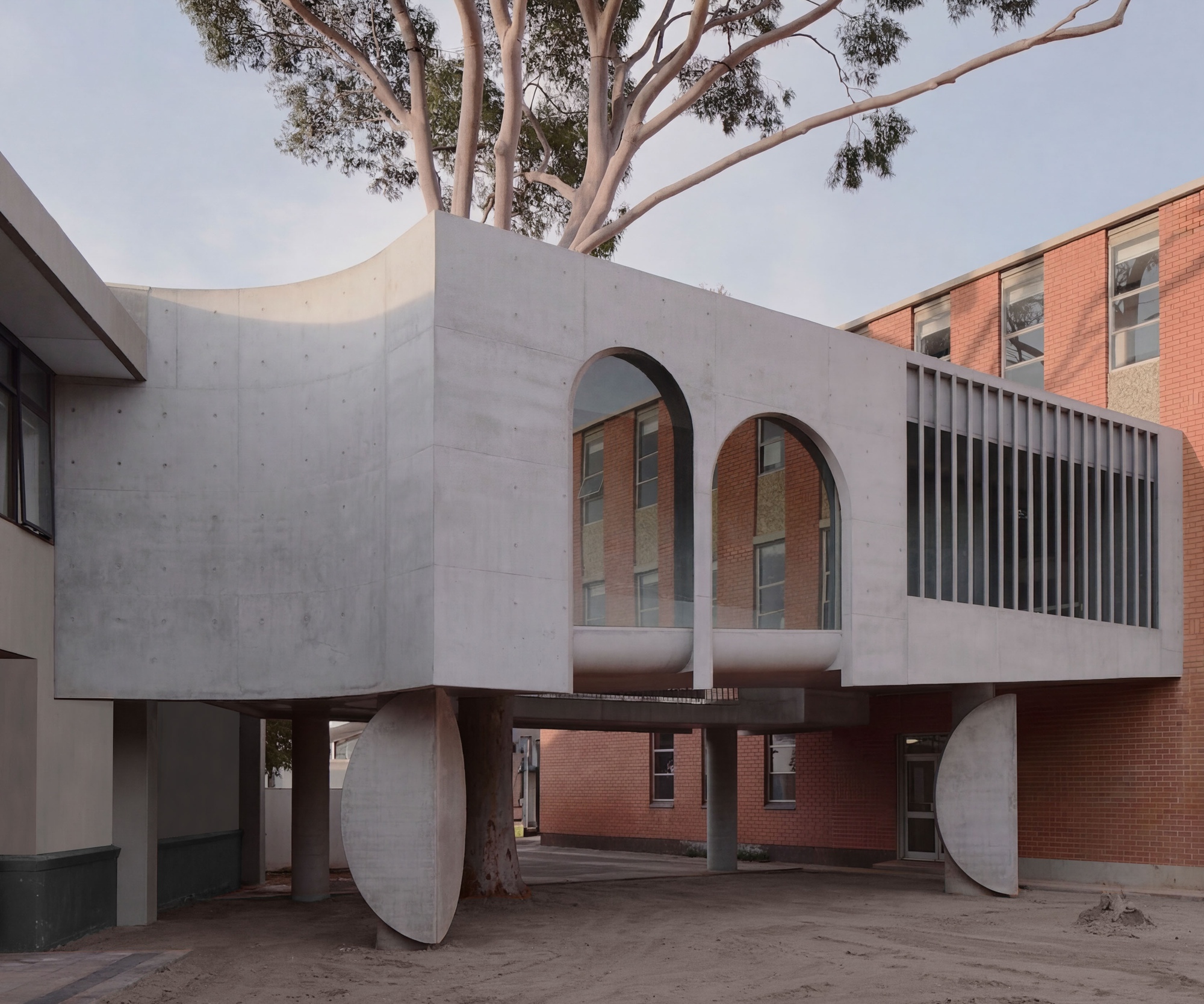 Elevated concrete volume on circular legs with arched openings framing an existing brick building