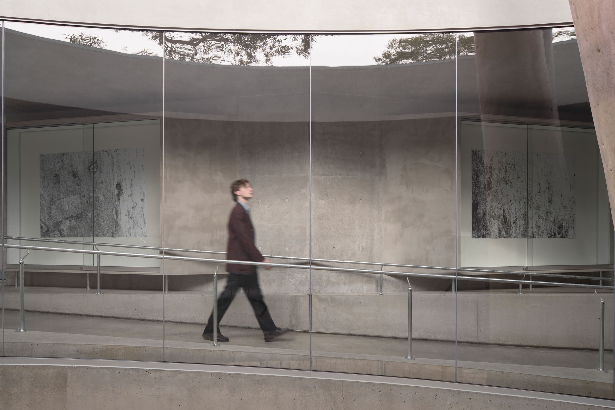 Glass-walled gallery corridor with a figure walking past concrete walls and horizontal metal railings