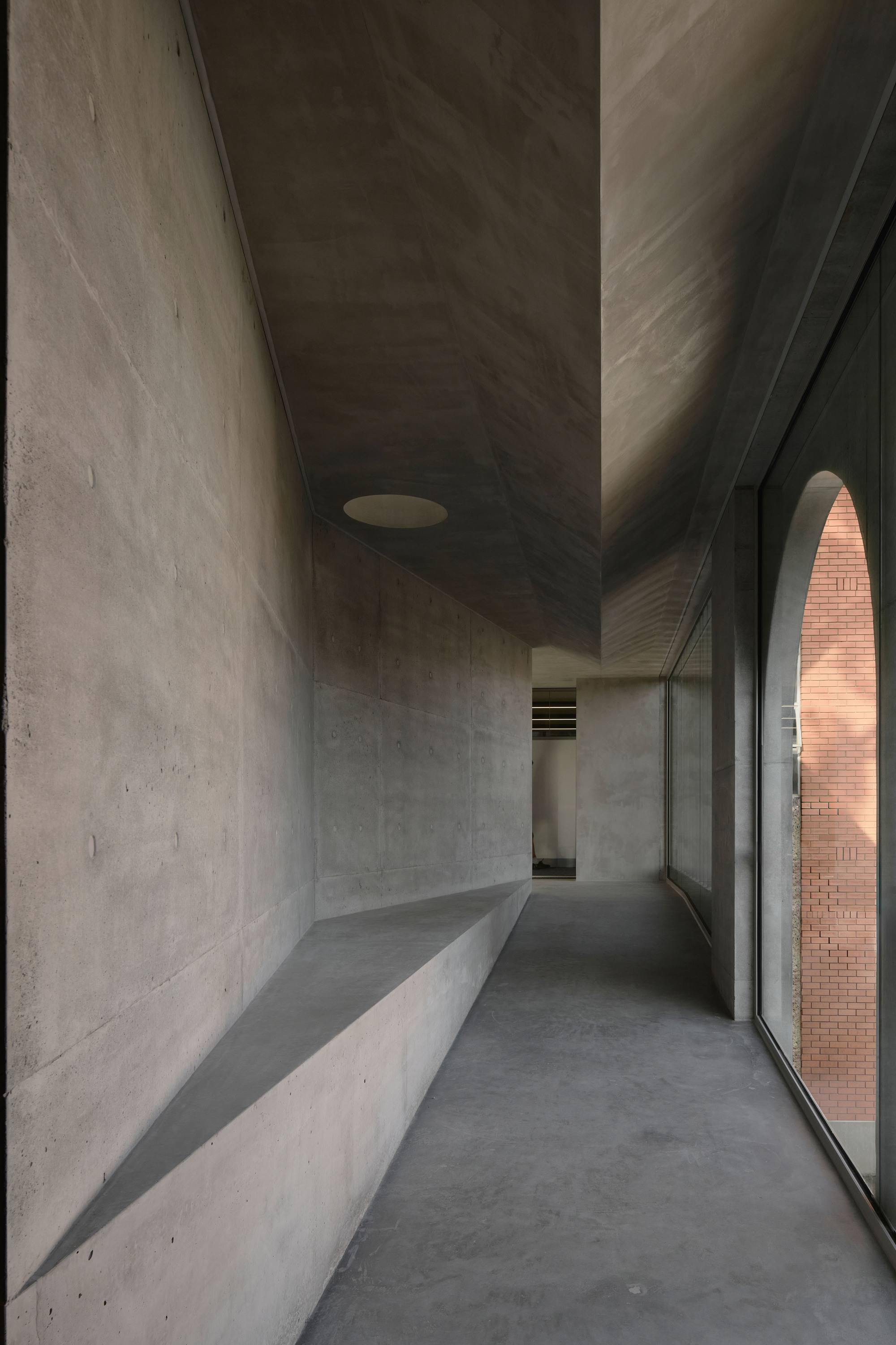 Concrete corridor with built-in bench along one wall and full-height glazing overlooking brick courtyard