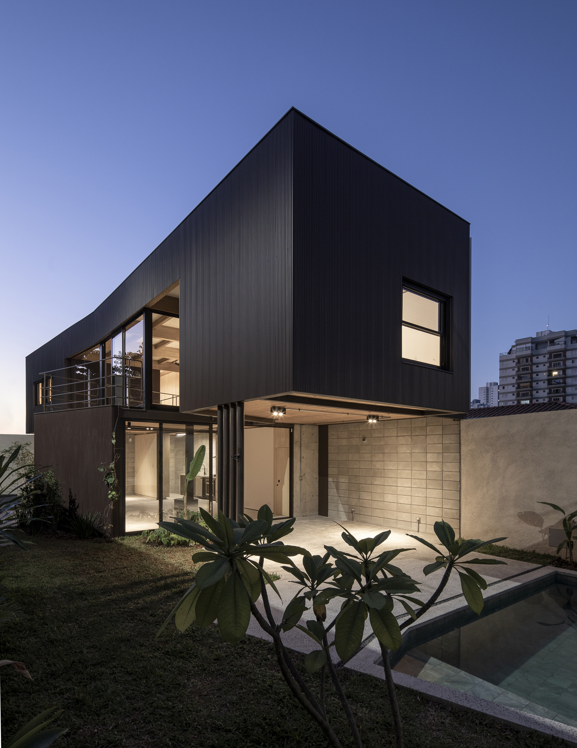 Dark corrugated facade with illuminated windows and cantilevered upper volume at dusk