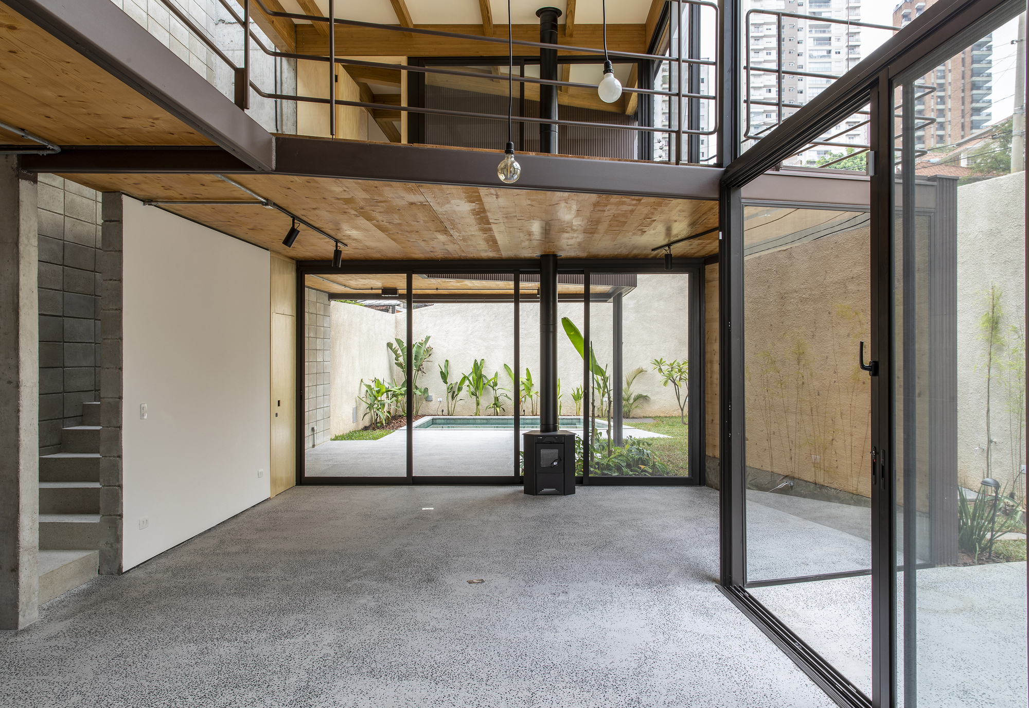 Double-height living space with plywood ceiling, steel mezzanine and glazed doors opening to a planted courtyard