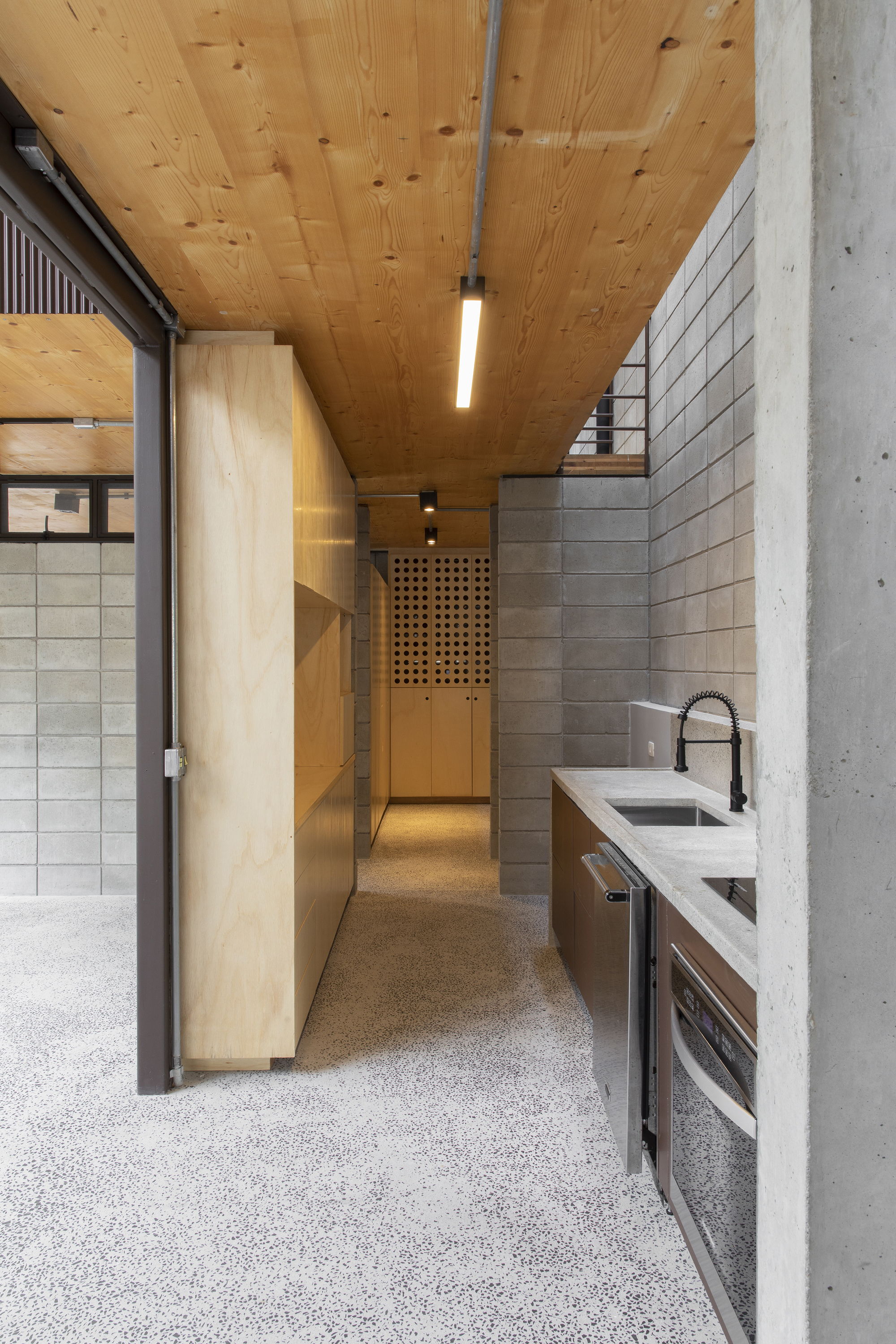 Galley kitchen with terrazzo flooring, plywood wardrobes and concrete block walls beneath a timber plank ceiling