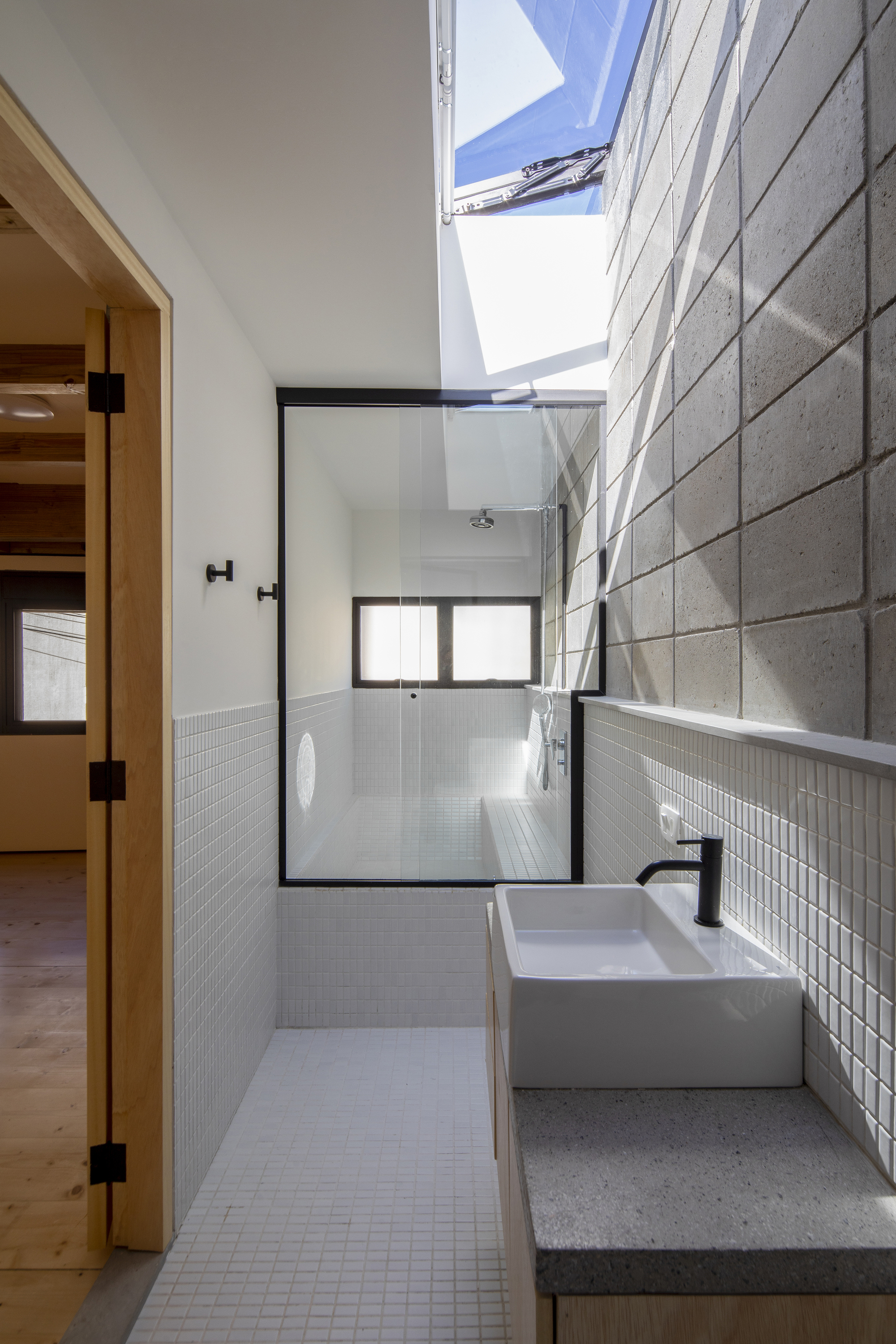 Bathroom with white mosaic tile walls, glass shower enclosure, concrete basin counter and overhead skylight